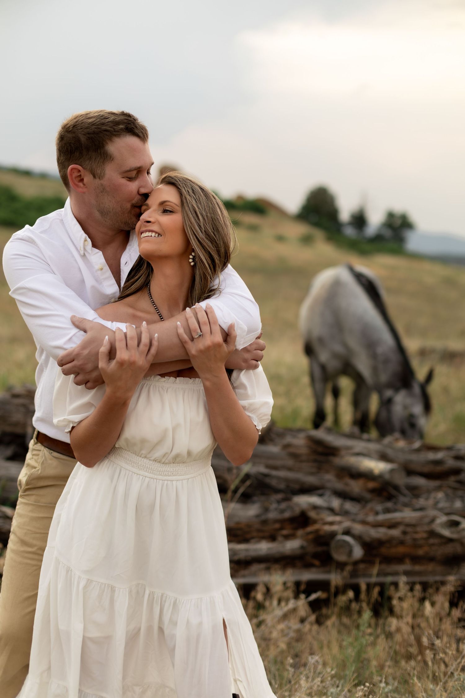 Couple embracing in a field with a gray horse grazing in the background during a romantic outdoor engagement photo session.