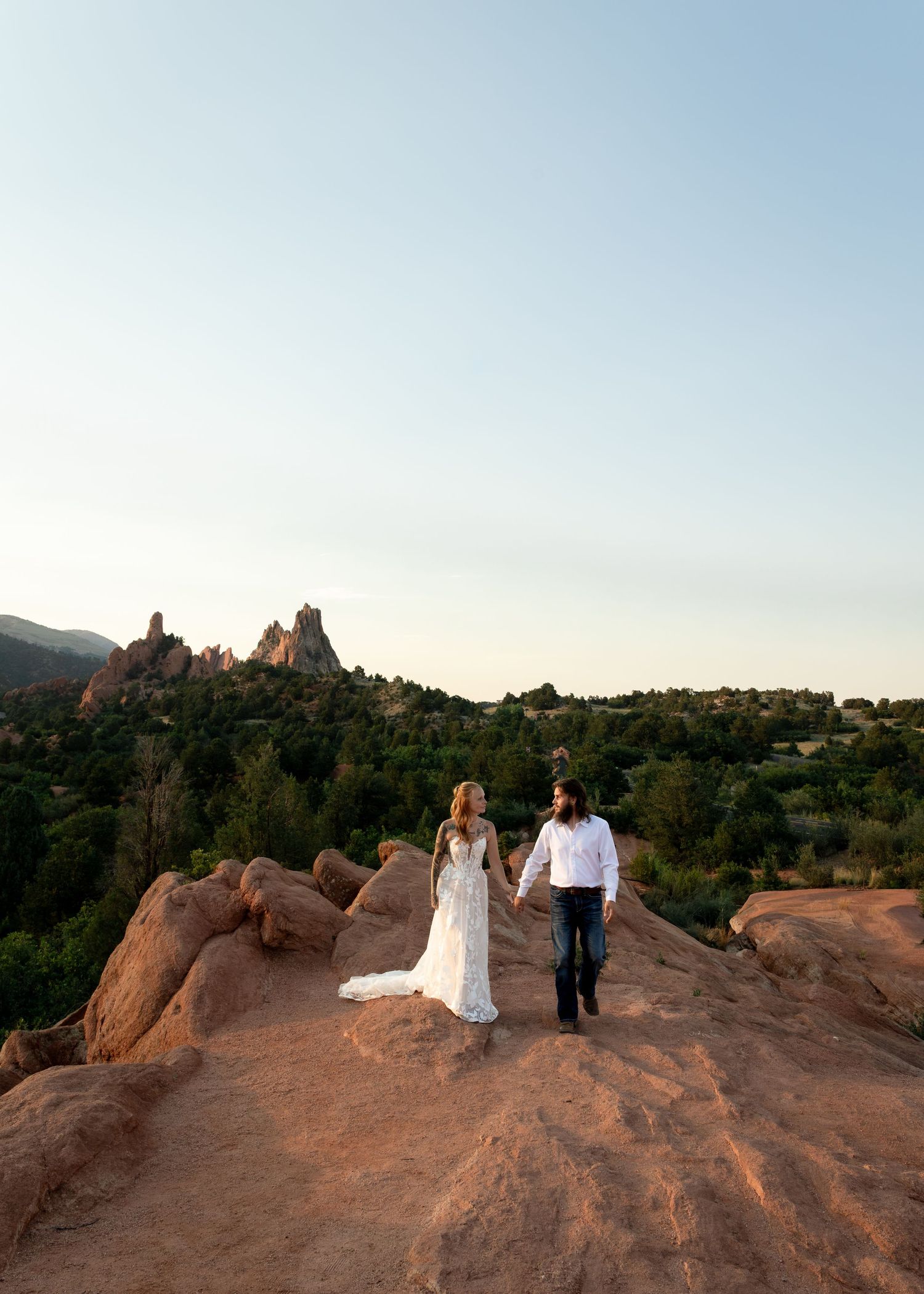 Couple in wedding attire hold hands on red rocks with Garden of the Gods formations at dusk.