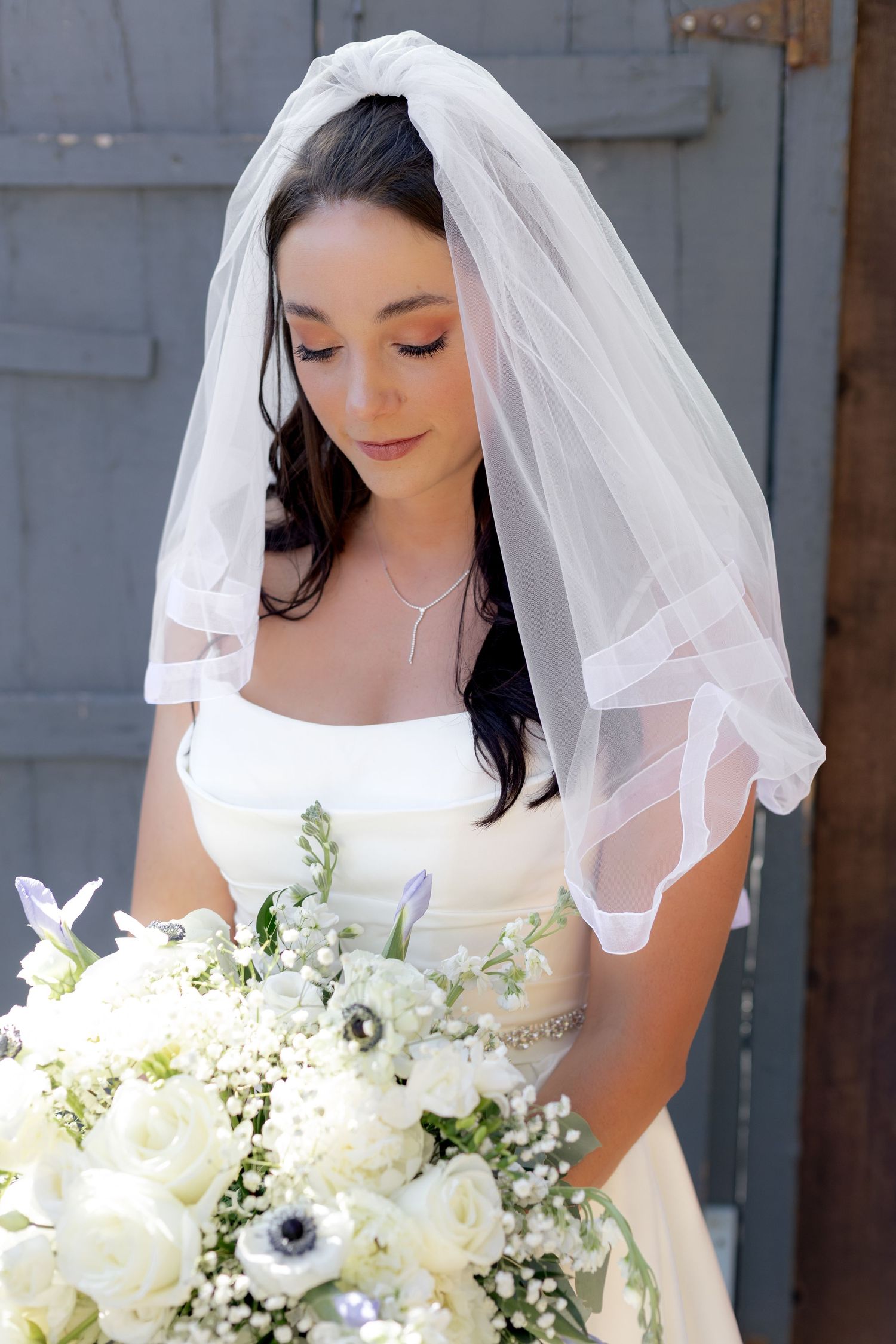 Beautiful bride wearing veil holding white rose bouquet with anemones on wedding day.