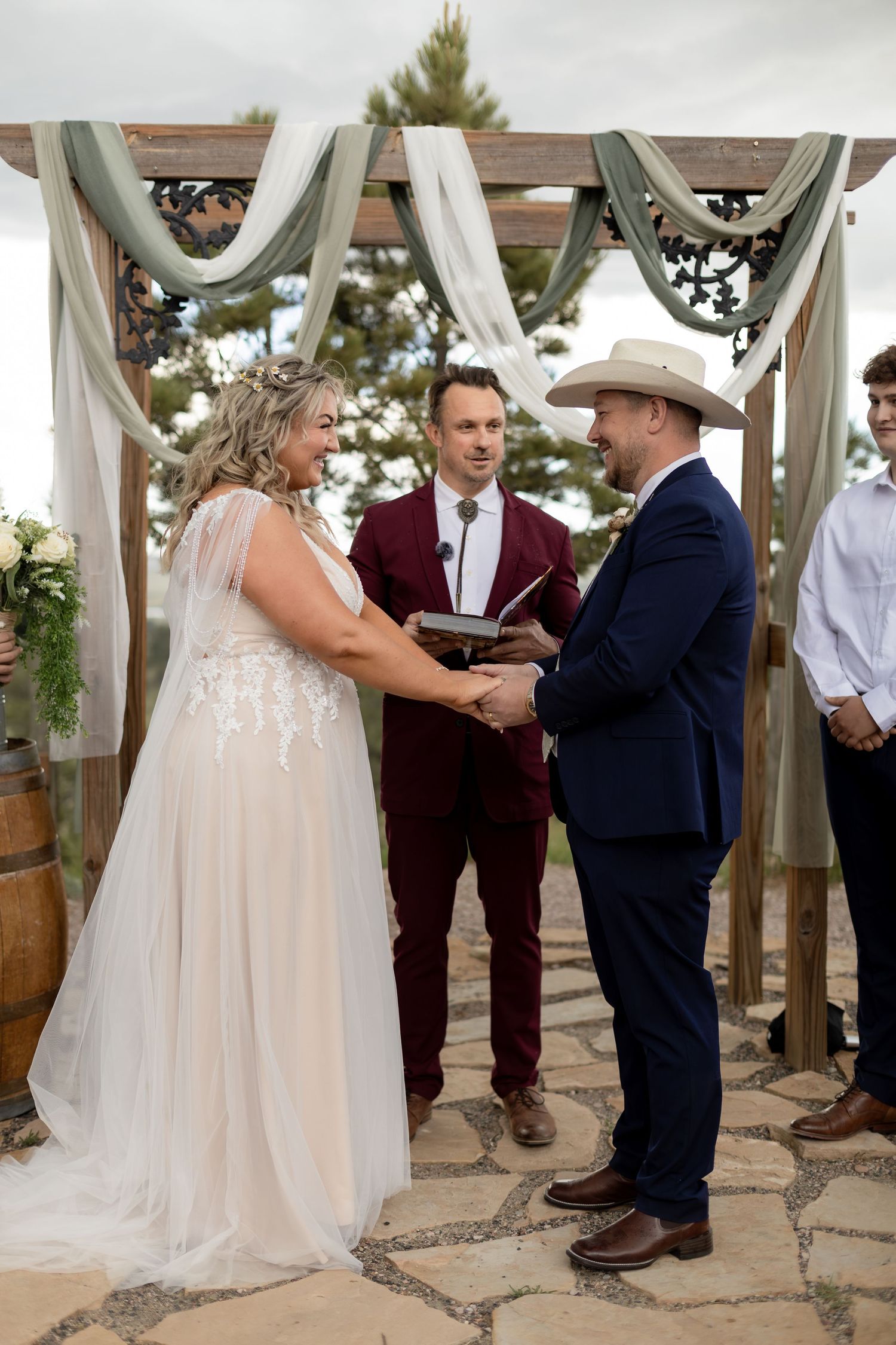 Bride and groom holding hands at rustic wooden arch altar during outdoor wedding ceremony.