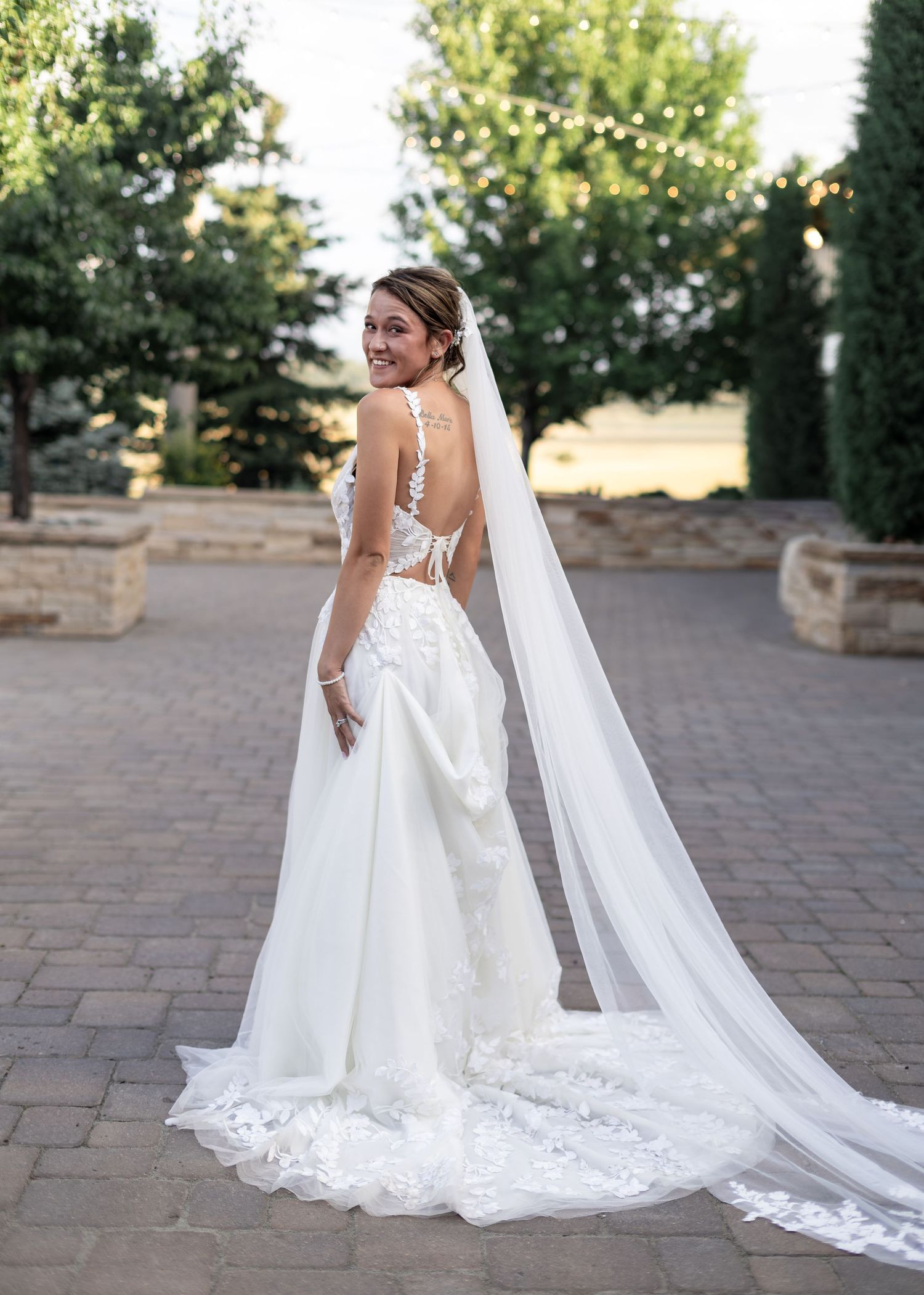 Bride in backless lace wedding gown with long veil on a paved outdoor venue at dusk.