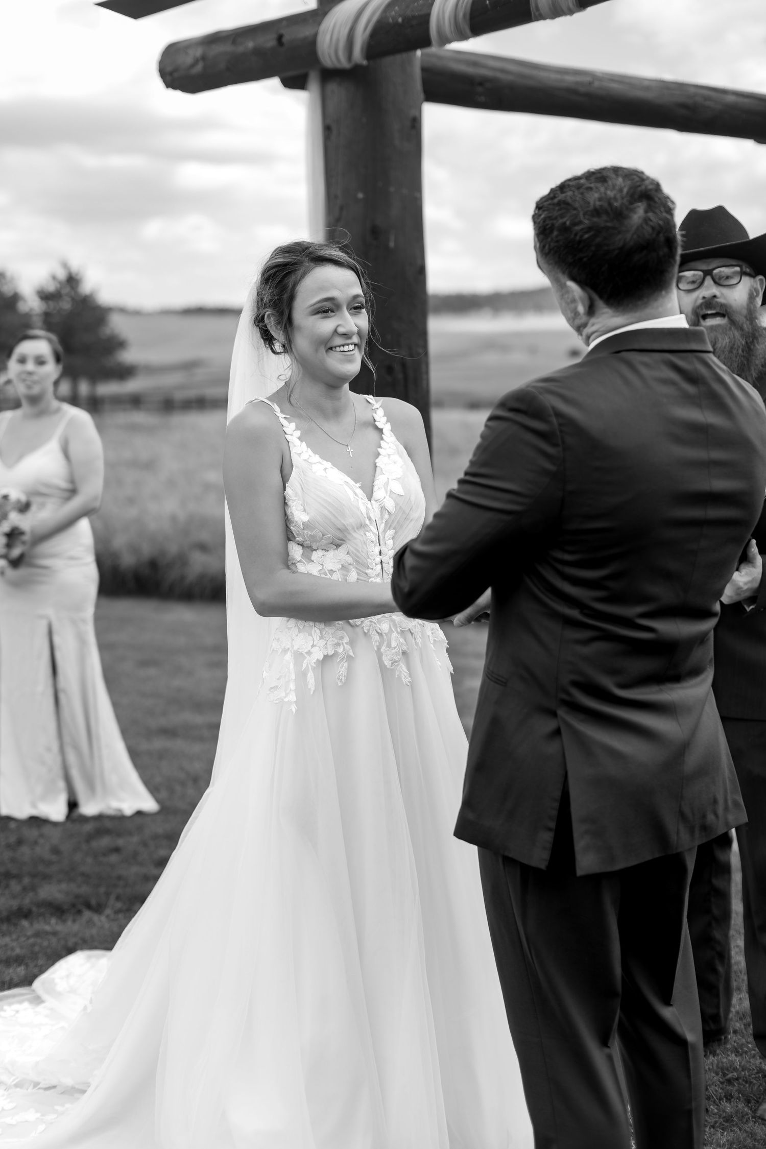 Black and white outdoor wedding ceremony with smiling bride in lace gown holding grooms hands before officiant.