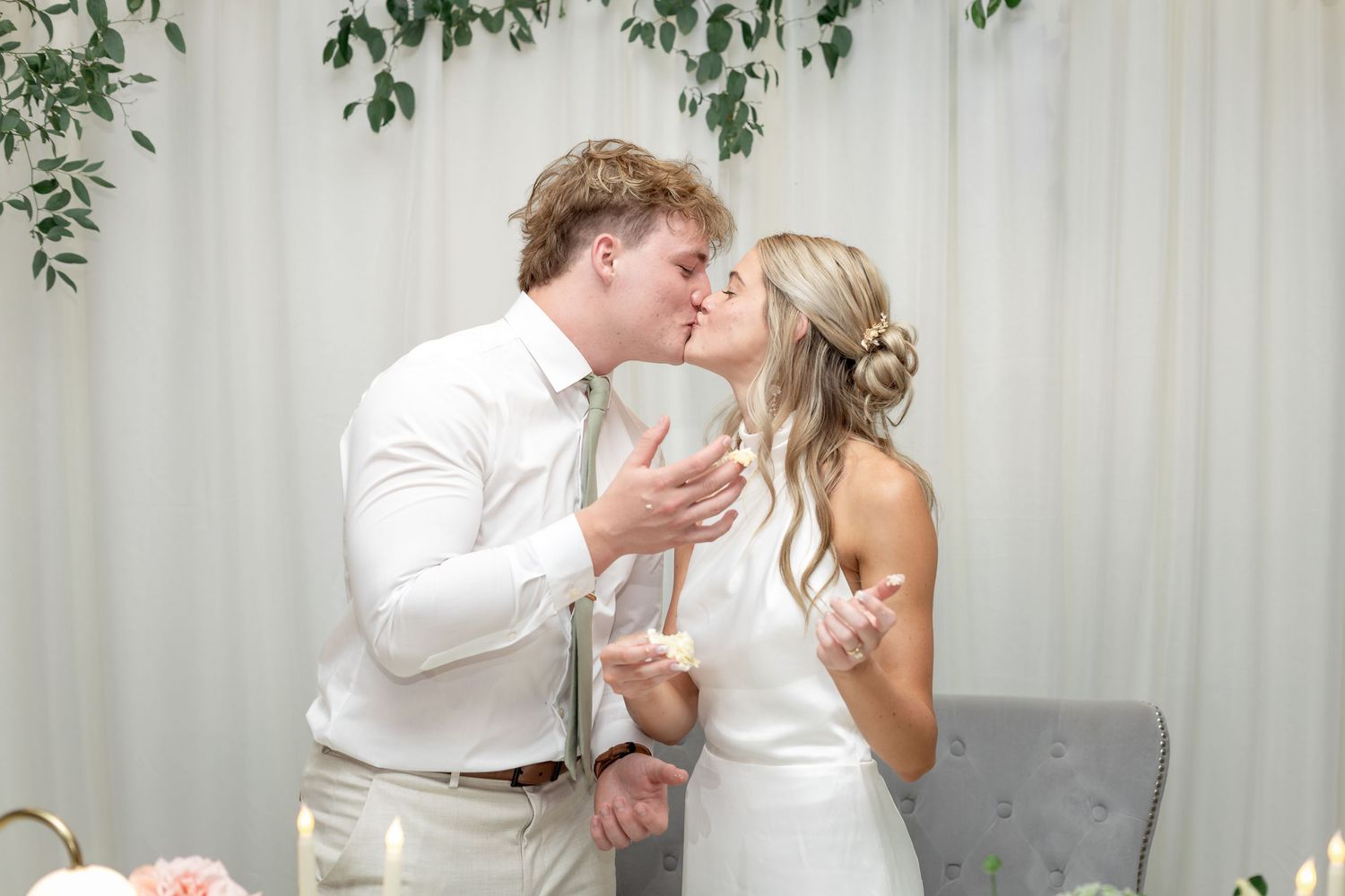 Bride and groom share a romantic kiss at their wedding reception, surrounded by floral decor and greenery backdrop.