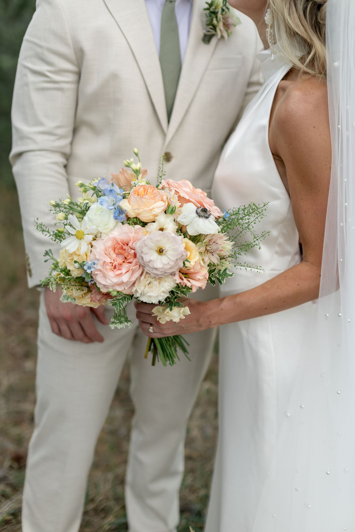 Bride holding a pastel bouquet of peach, white, and blue flowers beside groom in beige suit.