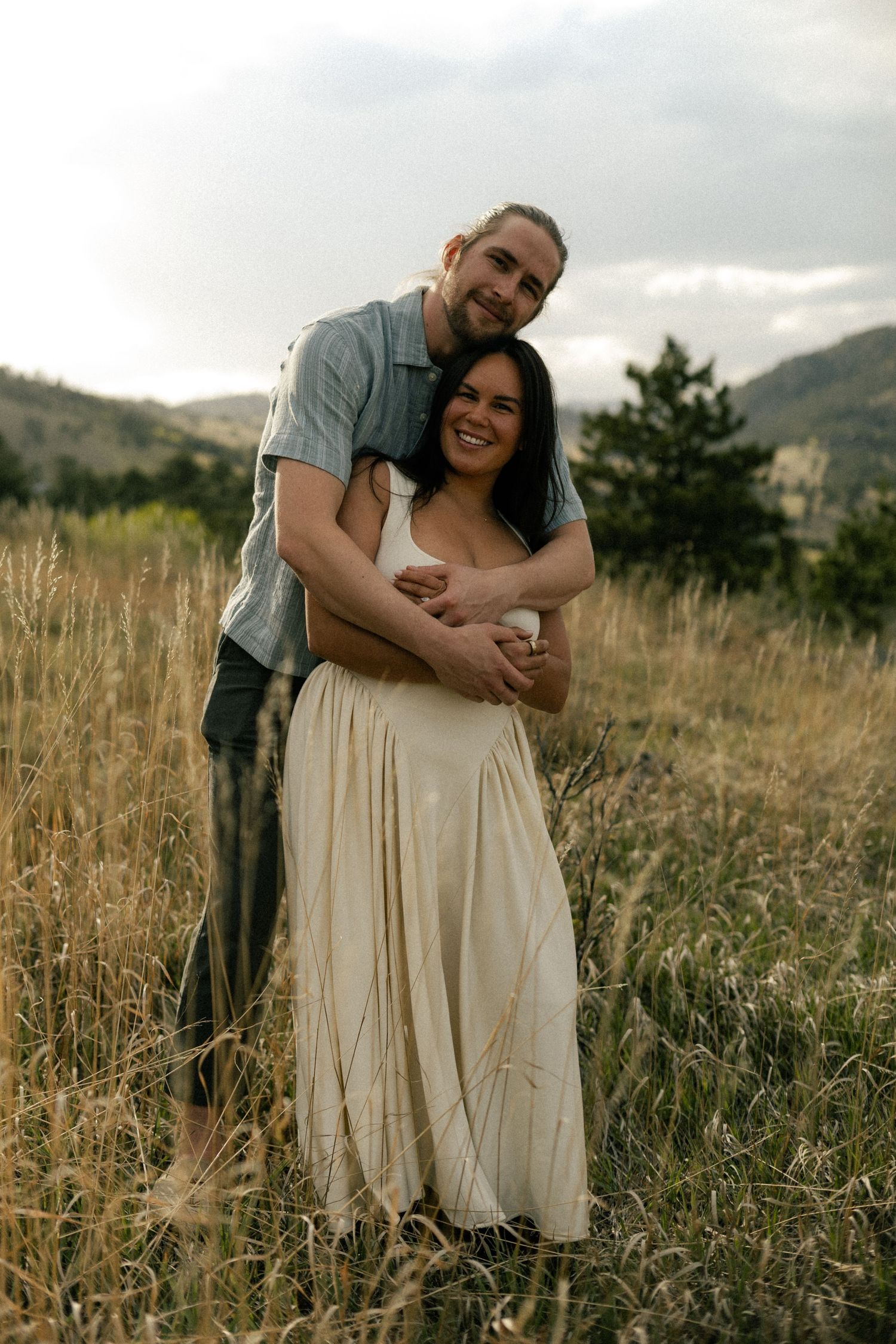Happy couple embracing in tall golden grass field with mountains, woman in flowing cream dress smiling.