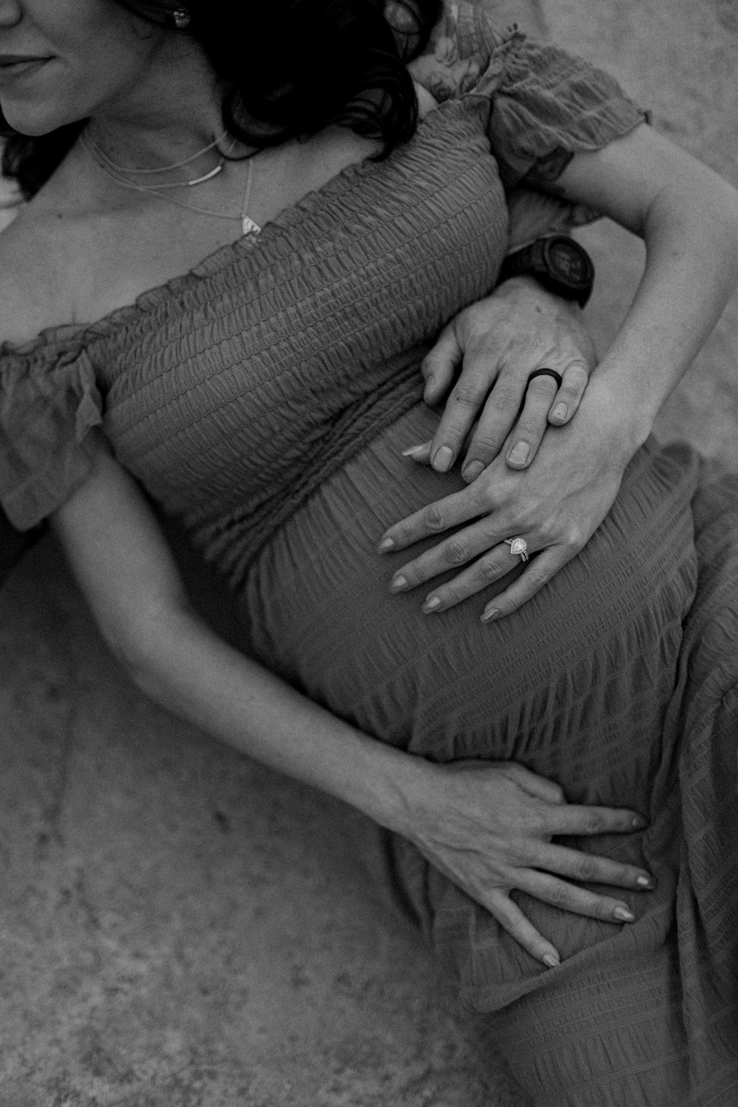 Black and white close-up of couple's hands gently holding pregnant belly, woman wearing engagement ring.
