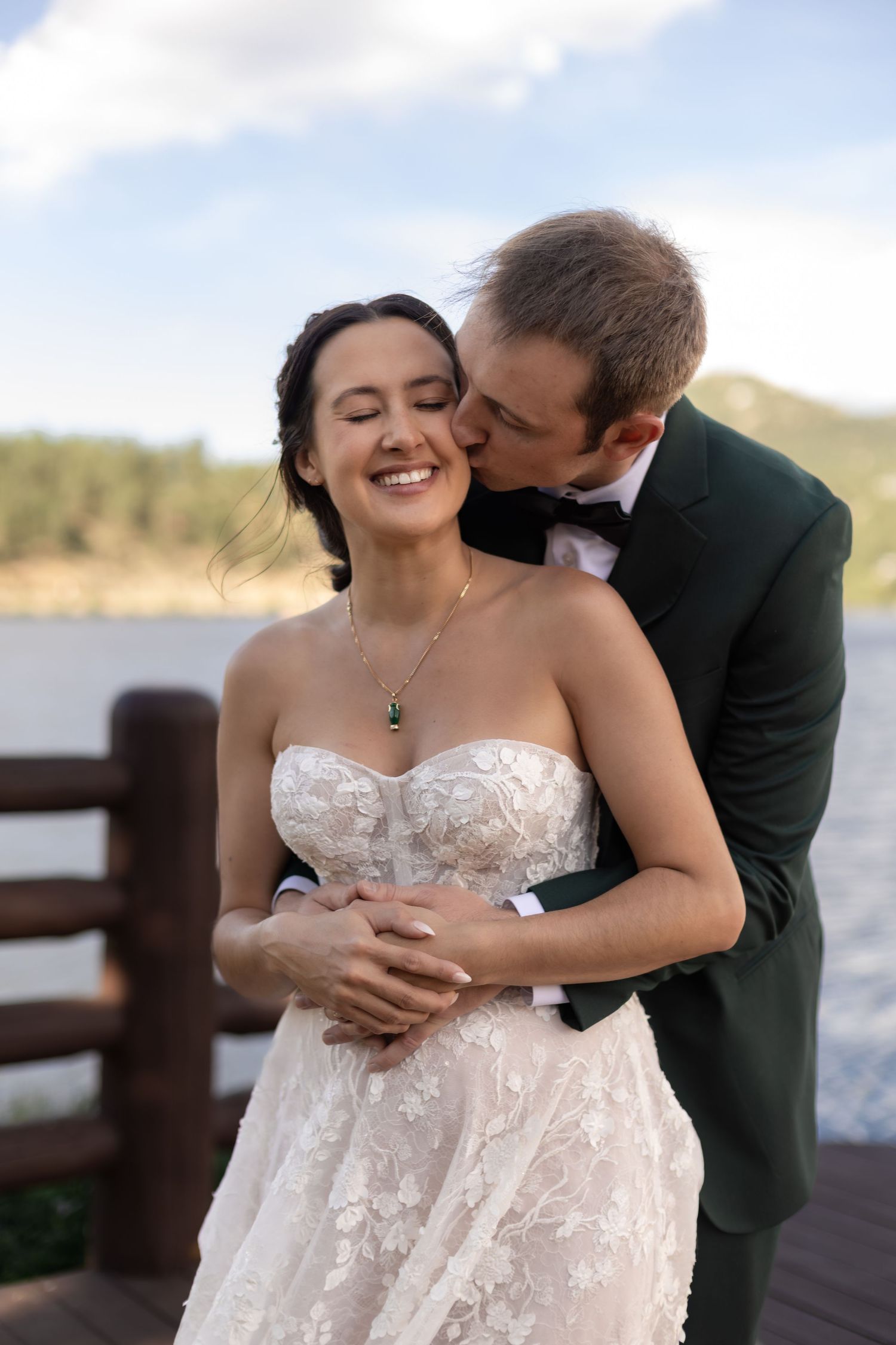 A groom in a dark green tuxedo kisses his smiling bride in a lace strapless gown on a lakeside dock.