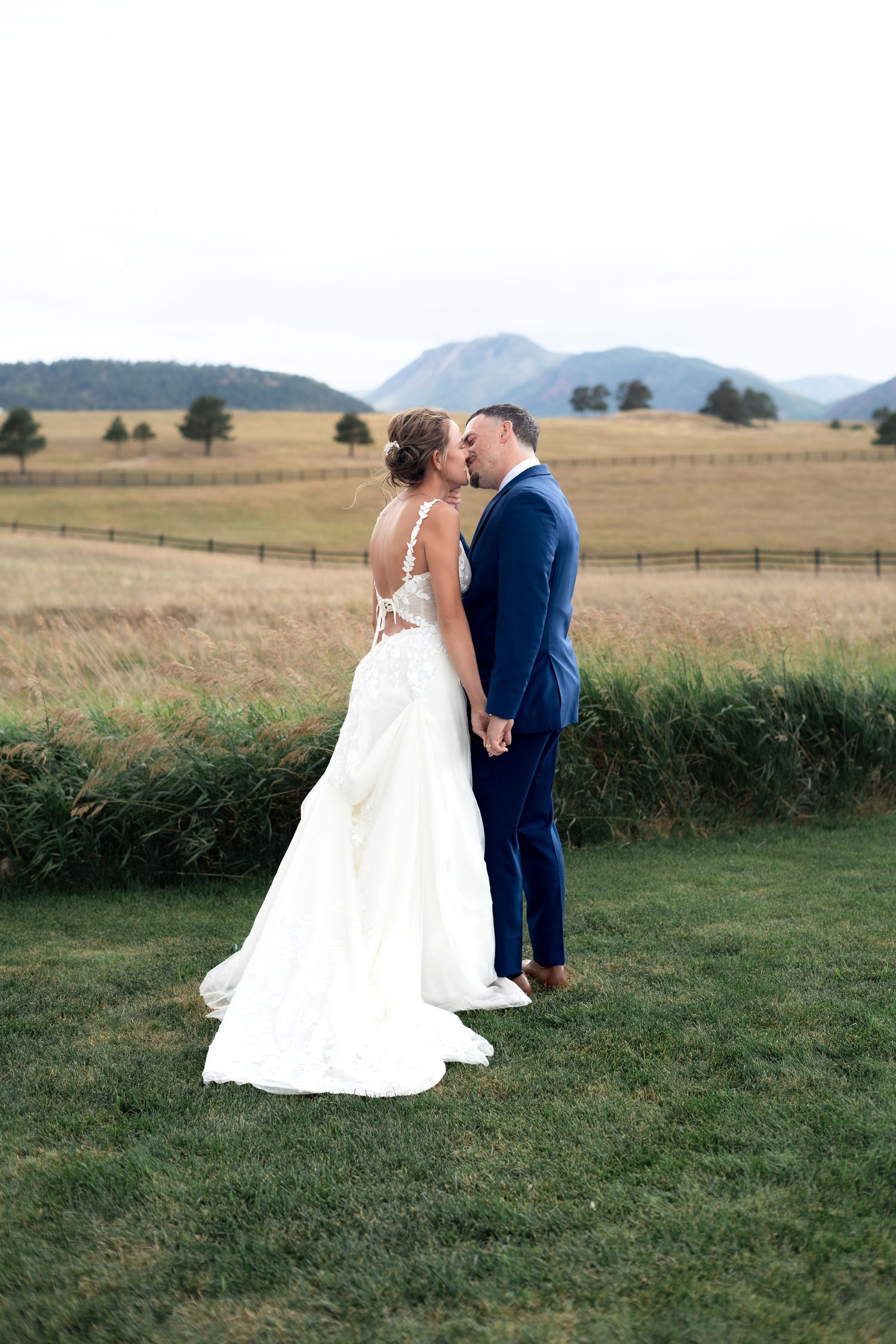 Wedding couple kissing on green lawn with golden fields and mountain range in background.