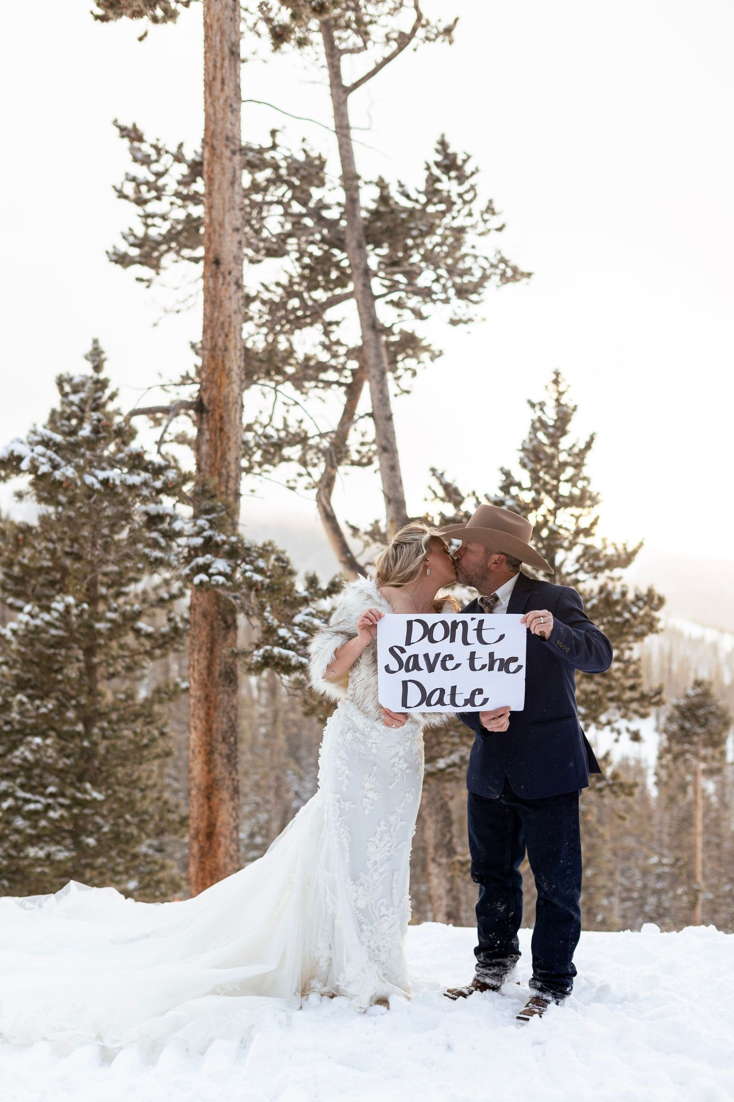Couple kissing in snow holding Don't Save the Date sign, winter wedding photo among snow-covered pine trees.