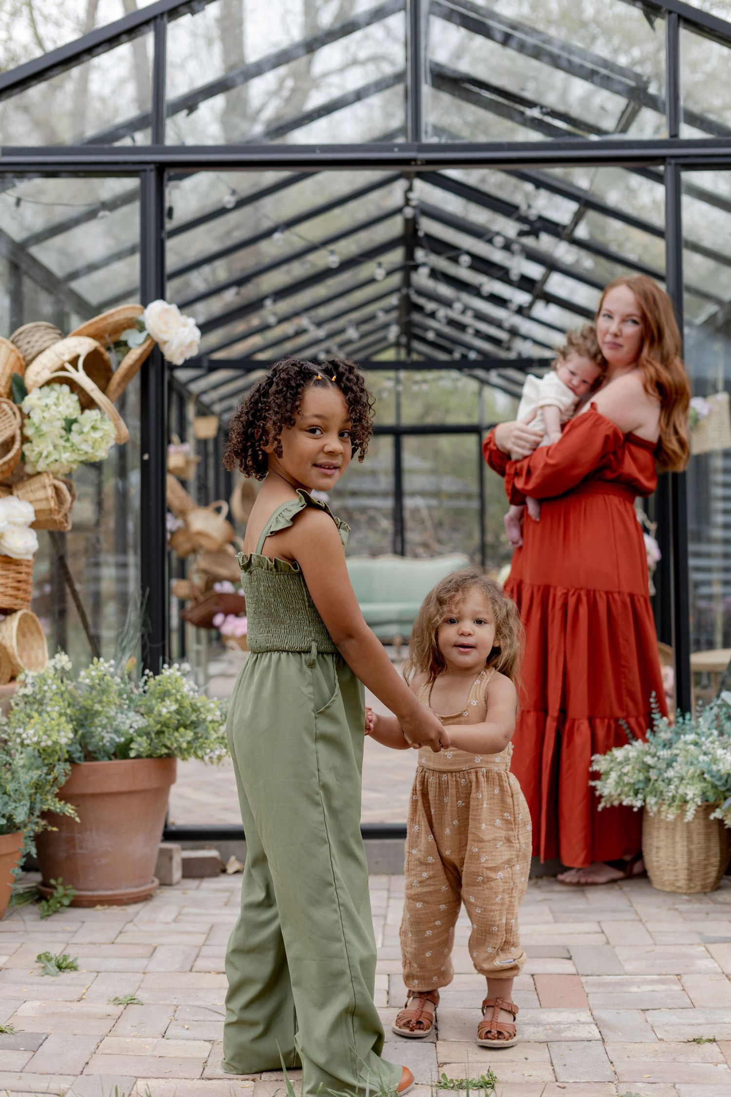 A woman in a red dress holds a baby outside a greenhouse while two young girls in green and tan outfits hold hands.