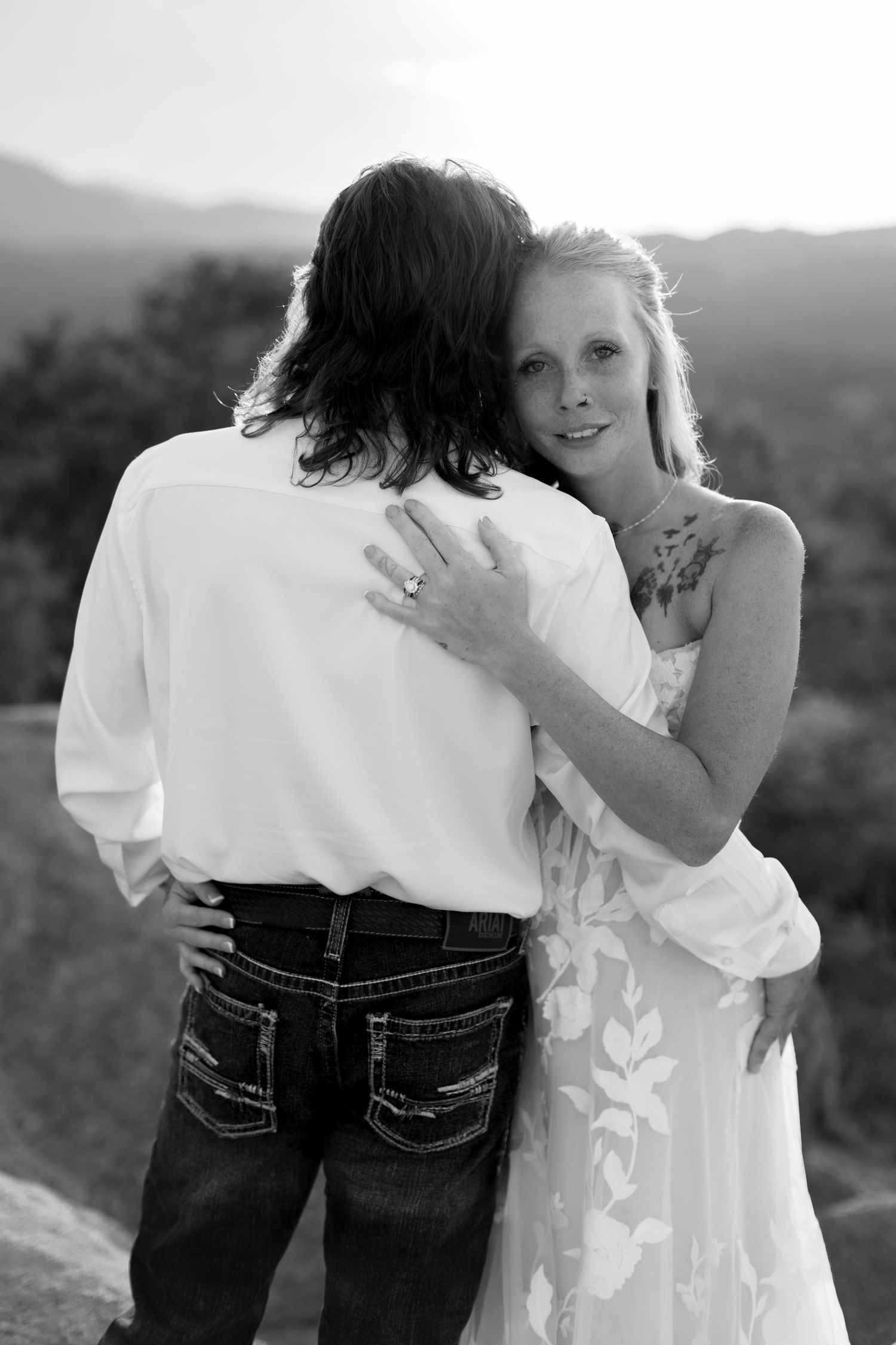 Black and white photo of bride hugging groom from behind at scenic mountain overlook.