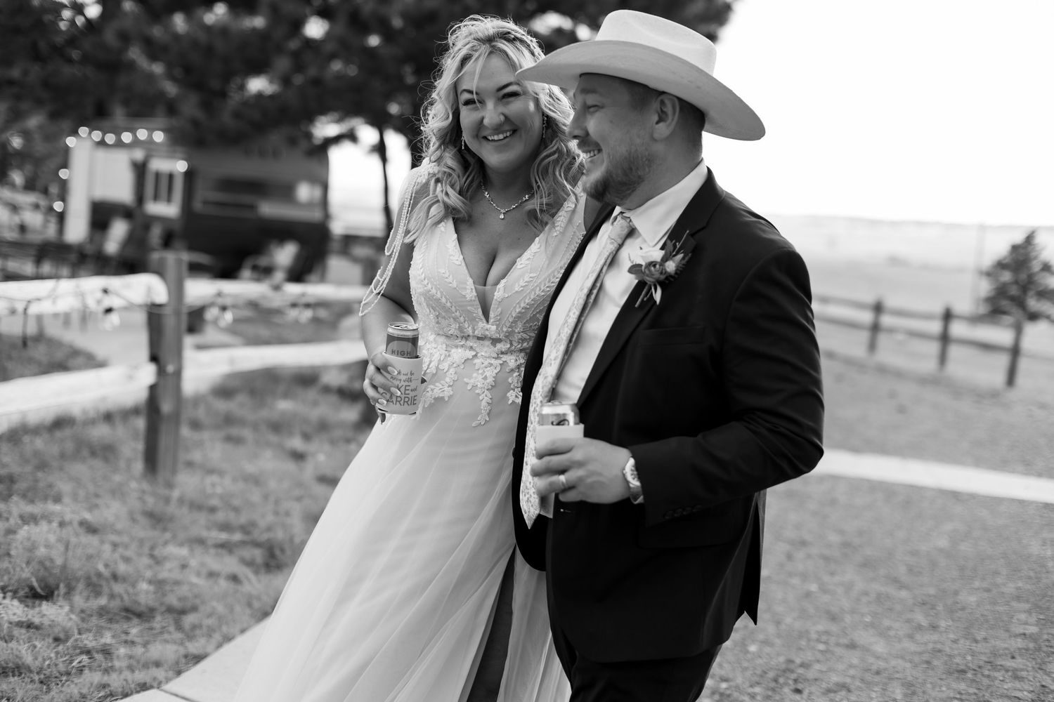 Smiling bride in white gown walks with groom in cowboy hat holding drinks at outdoor wedding reception.