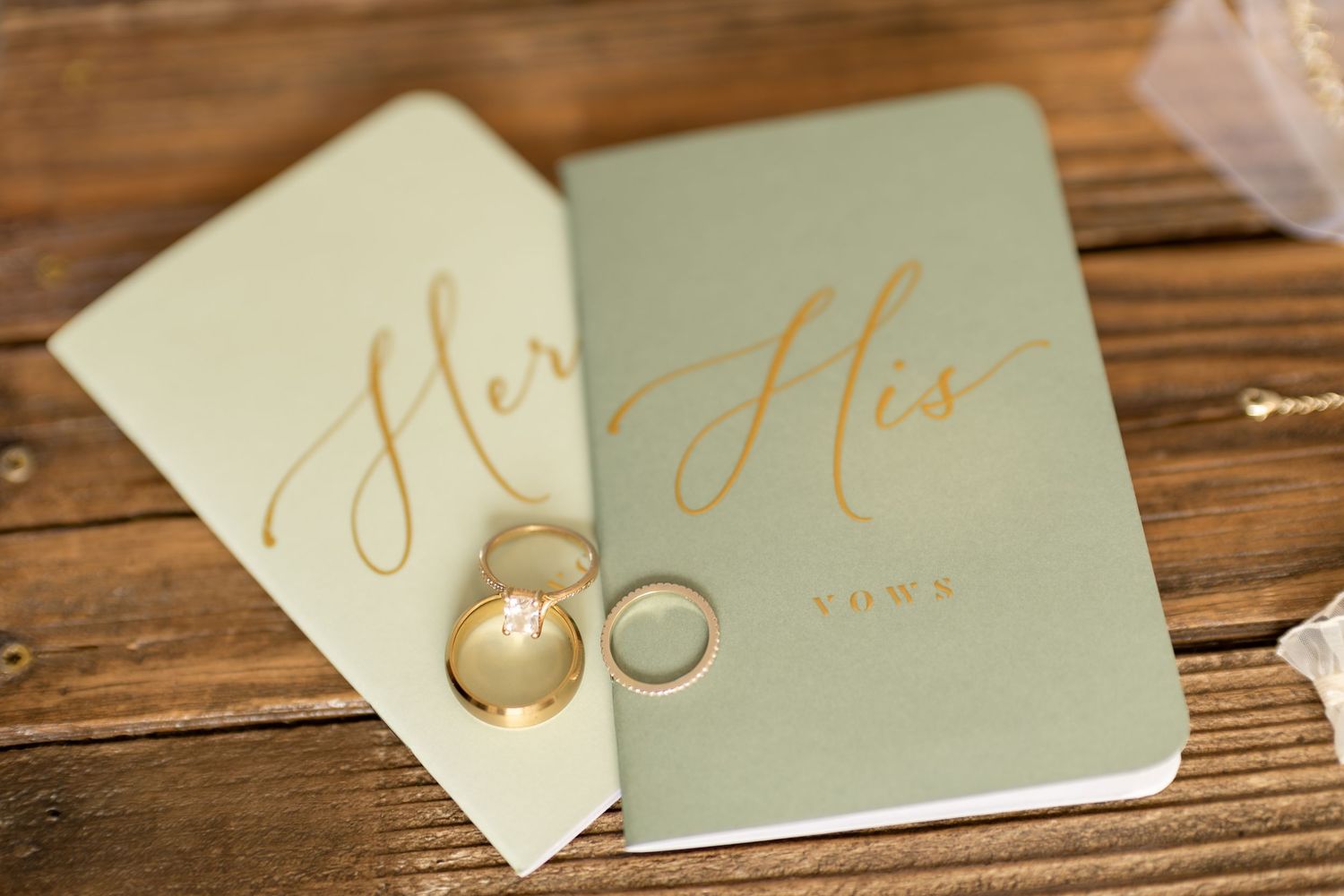His and Hers sage green vow books with gold calligraphy and wedding rings on rustic wooden surface.