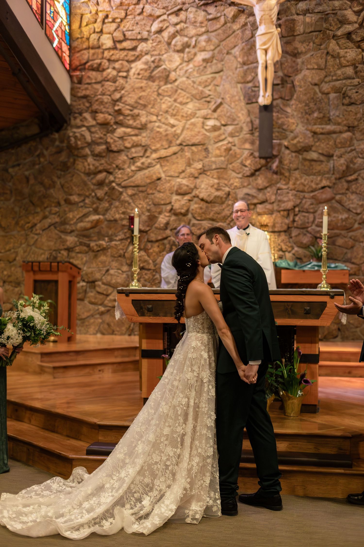 Bride and groom share first kiss at church altar during wedding ceremony with priest and crucifix in background.