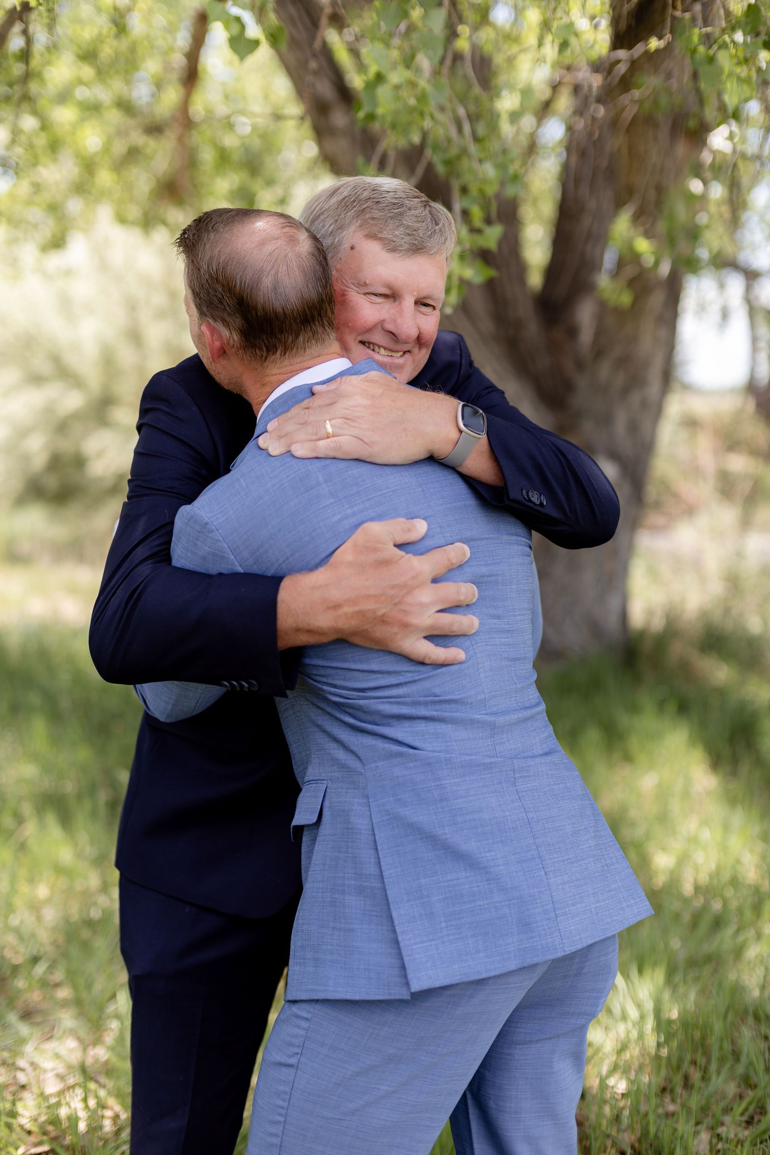 Two men in suits share a warm embrace outdoors under a tree at a wedding celebration.