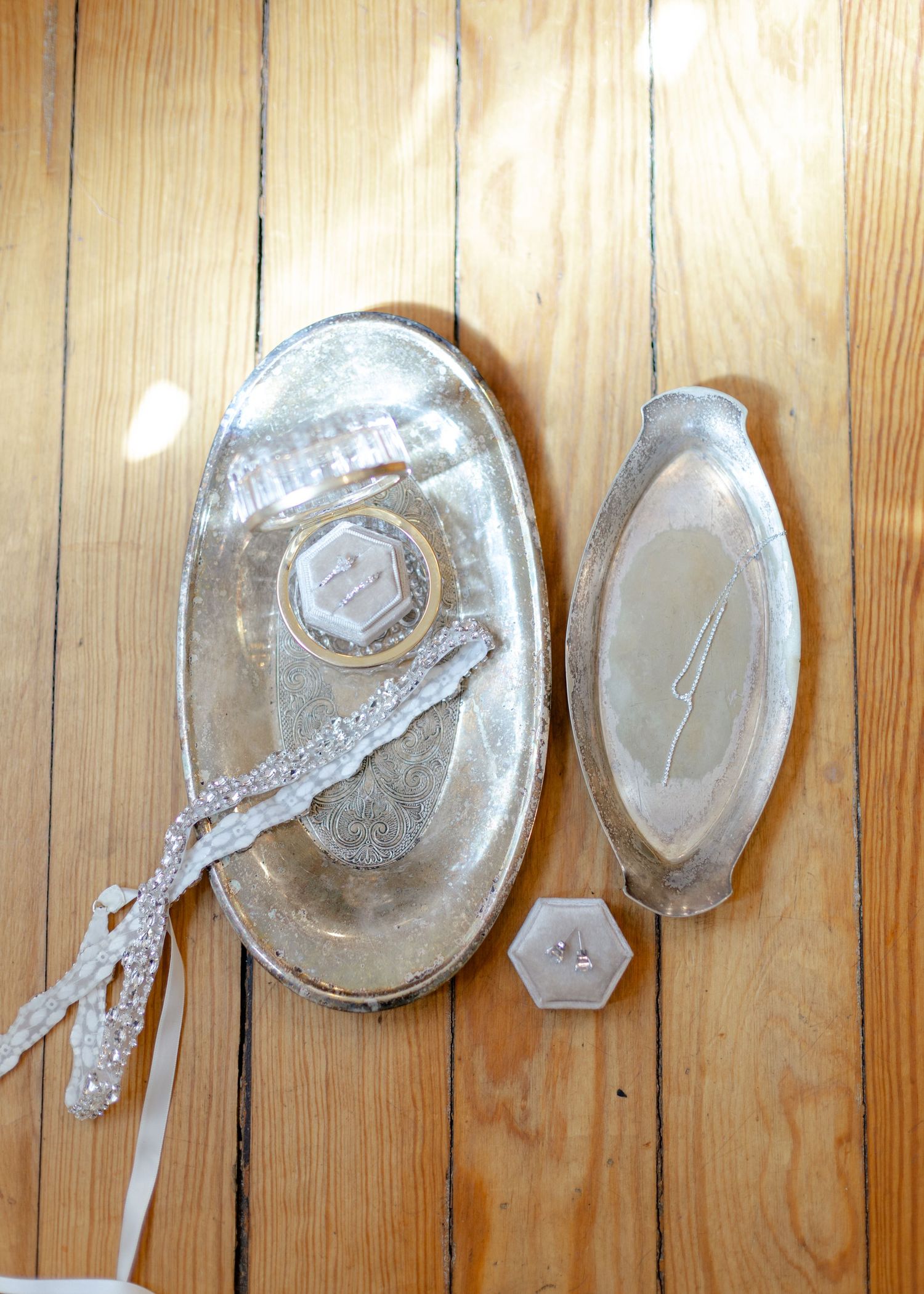 Silver oval wedding trays with ring box, lace ribbon, and small pink gems displayed on wooden floor.