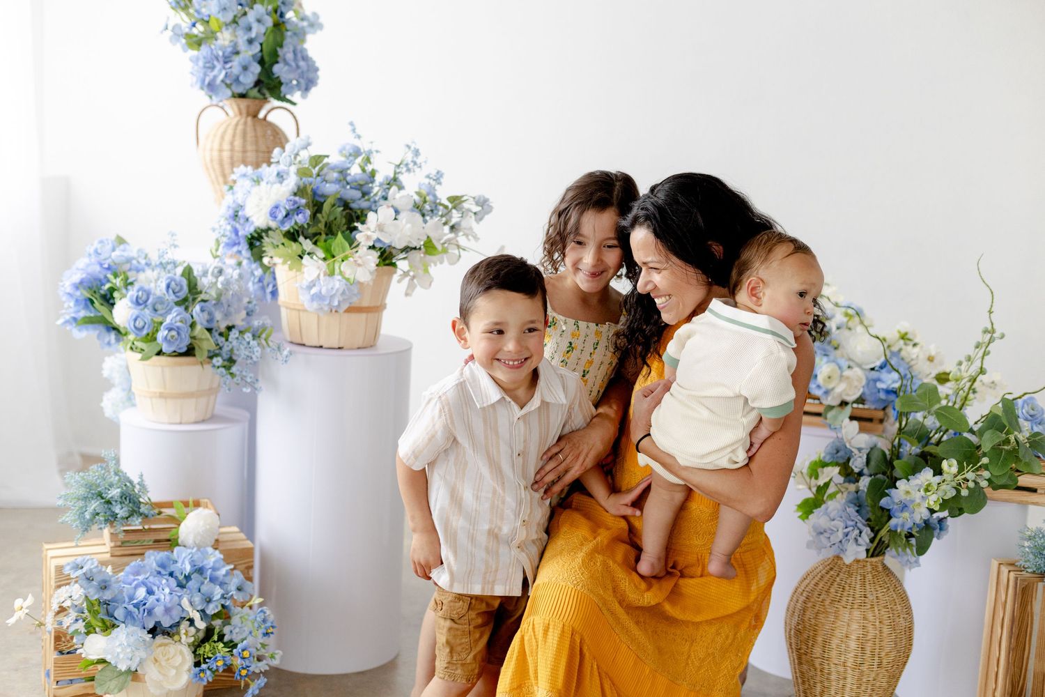 A mother in a yellow dress poses with her three children surrounded by blue hydrangea floral arrangements.