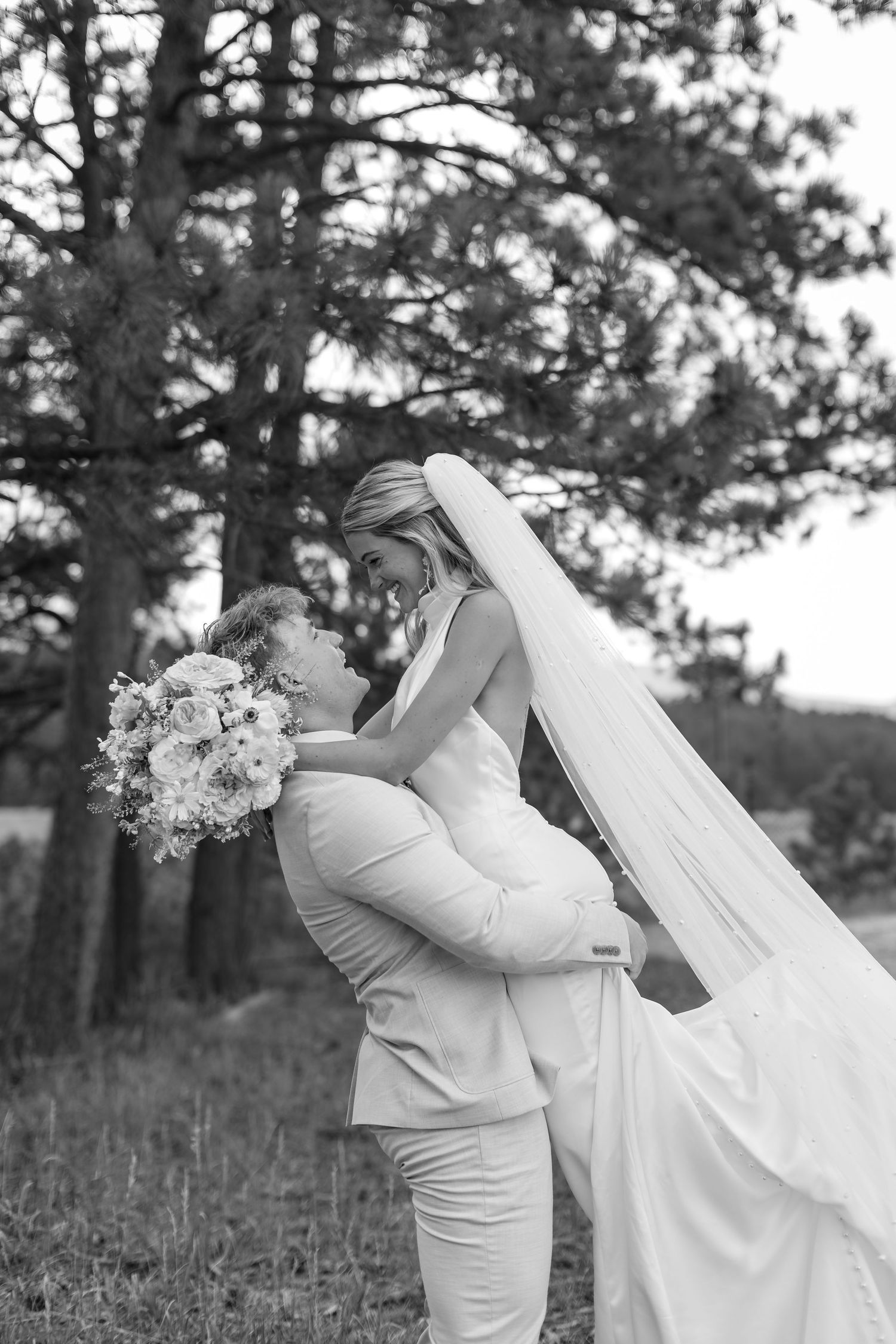 Black and white photo of groom lifting bride in wedding dress and veil, holding bouquet, surrounded by pine trees.