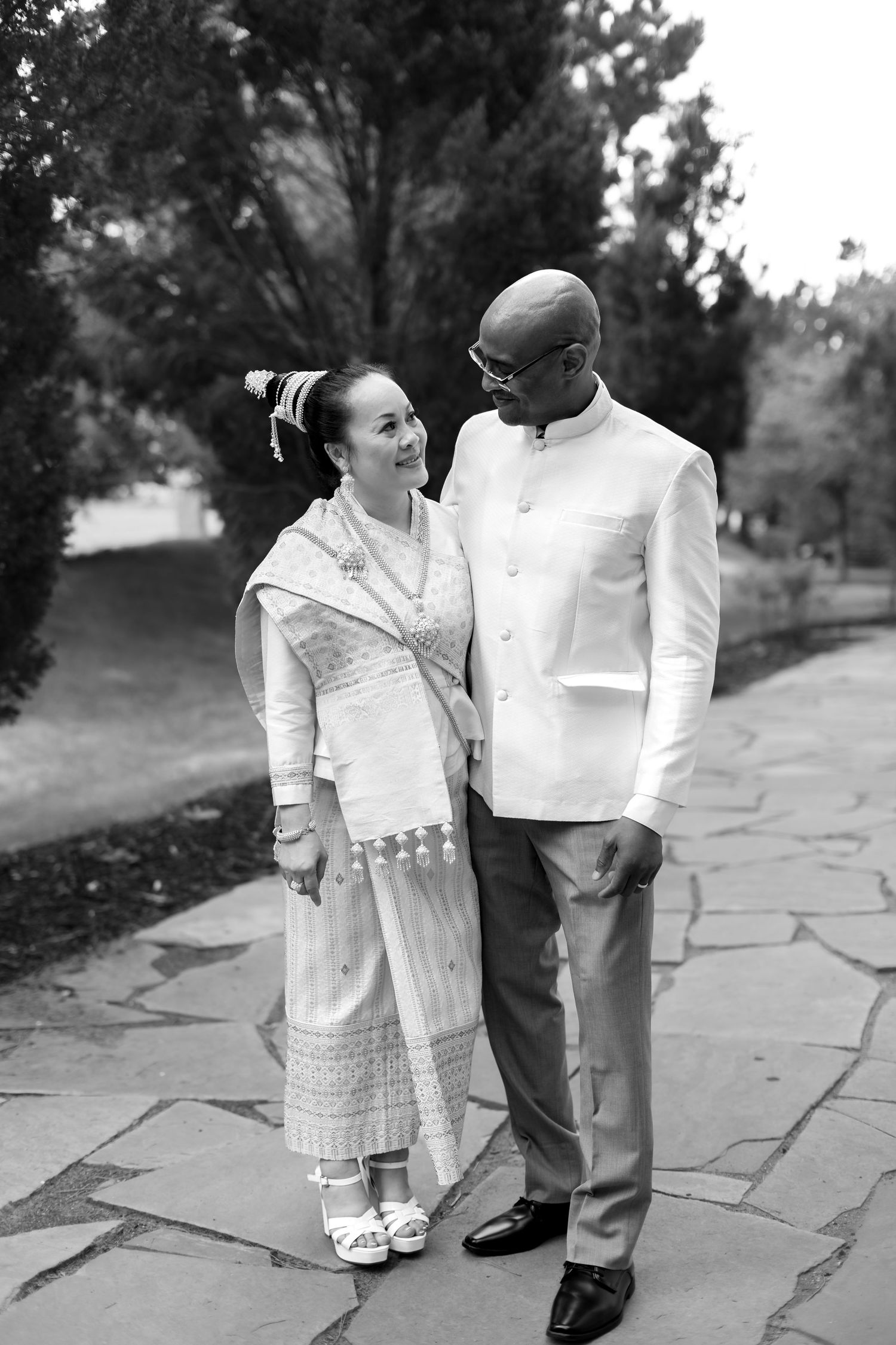 Black and white photo of an interracial couple in traditional cultural wedding attire standing on a stone path outdoors.