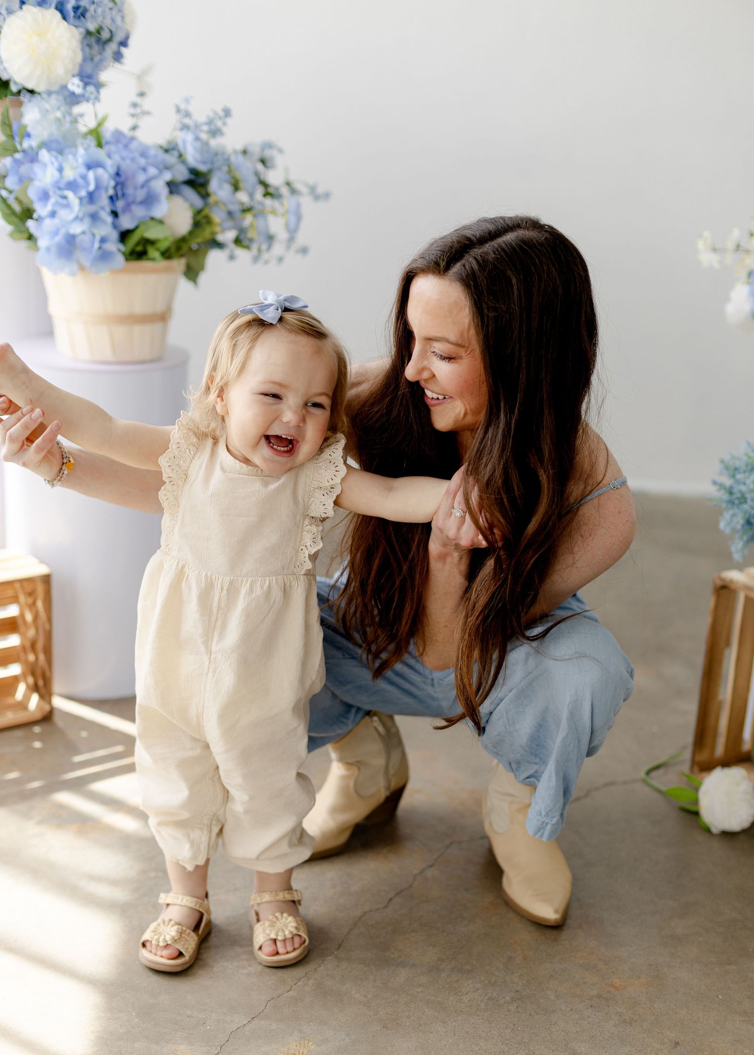 A smiling mother crouches beside her laughing toddler girl among blue hydrangea arrangements in a bright studio.