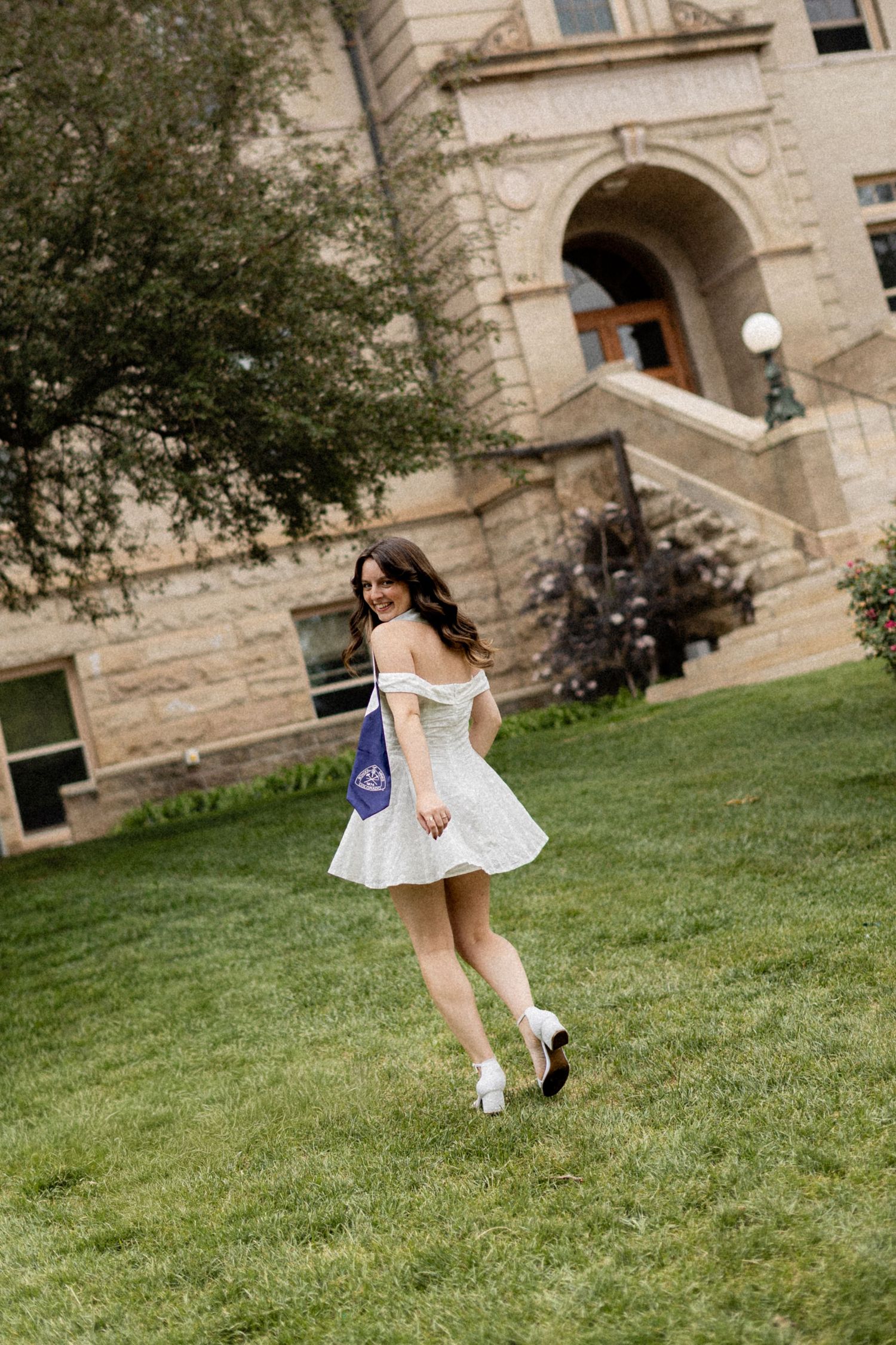 Young woman in white off-shoulder dress walks on green campus lawn near stone university building.