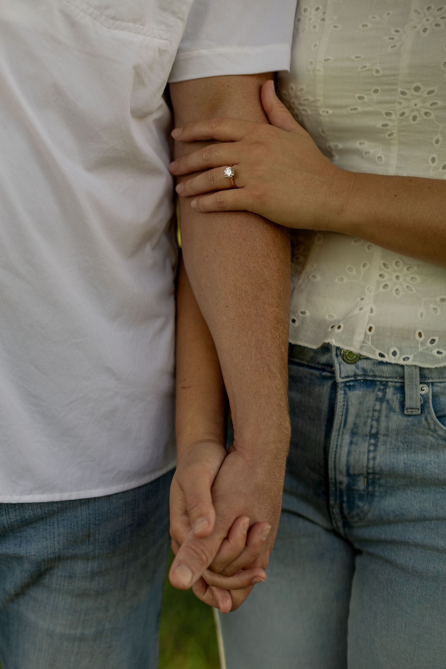 A couple holds hands tenderly, the woman wearing an engagement ring, both dressed casually in jeans and white tops.