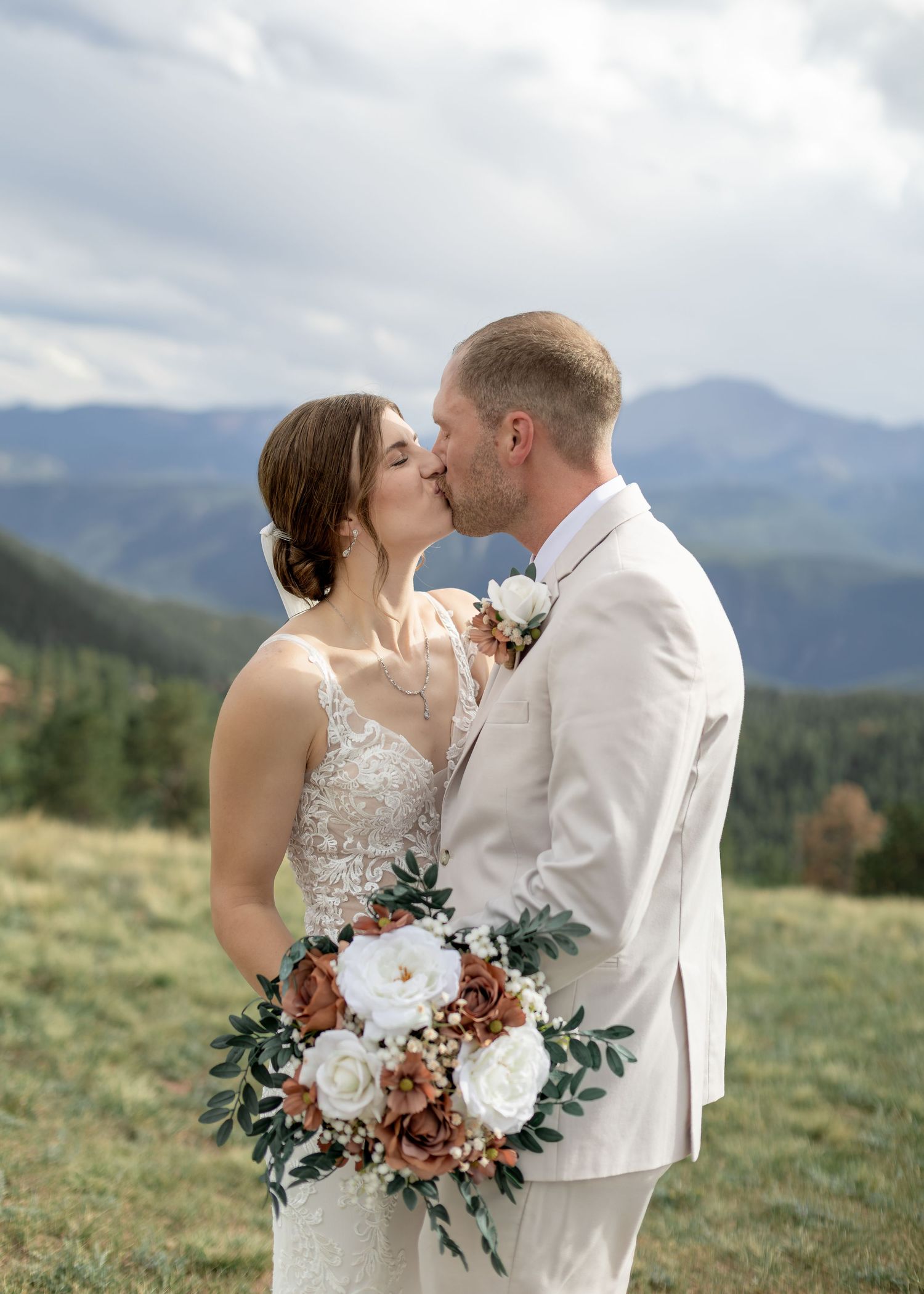 Bride and groom kissing on a mountain meadow, bride holding rustic bouquet of white and terracotta flowers with greenery.