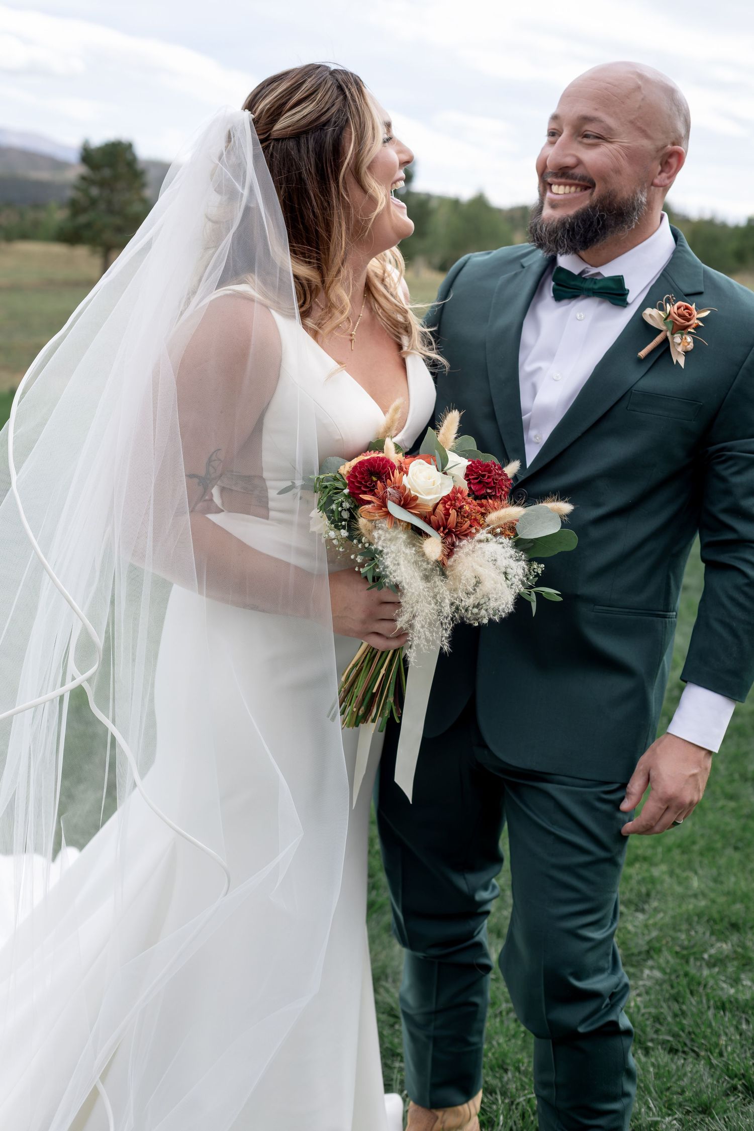 Smiling bride in white gown with veil holds floral bouquet beside groom in green suit with bow tie outdoors.