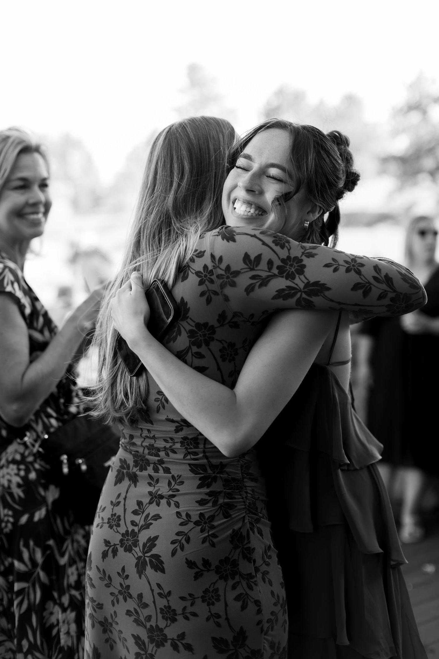 Two women share a warm embrace, smiling joyfully at an outdoor event, captured in black and white.