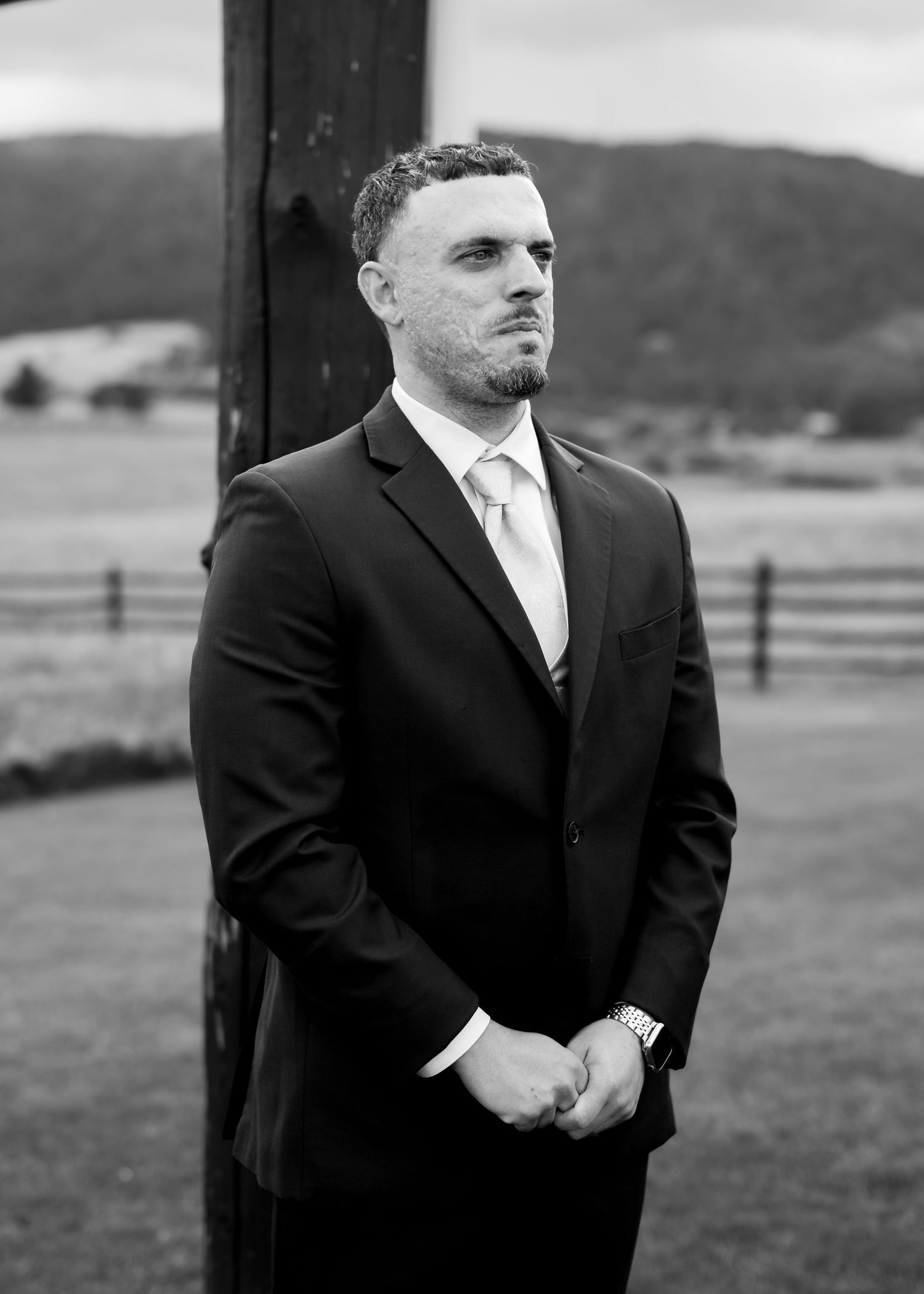 A man in a dark suit and light tie stands outdoors at a rural wedding venue in a black and white photo.
