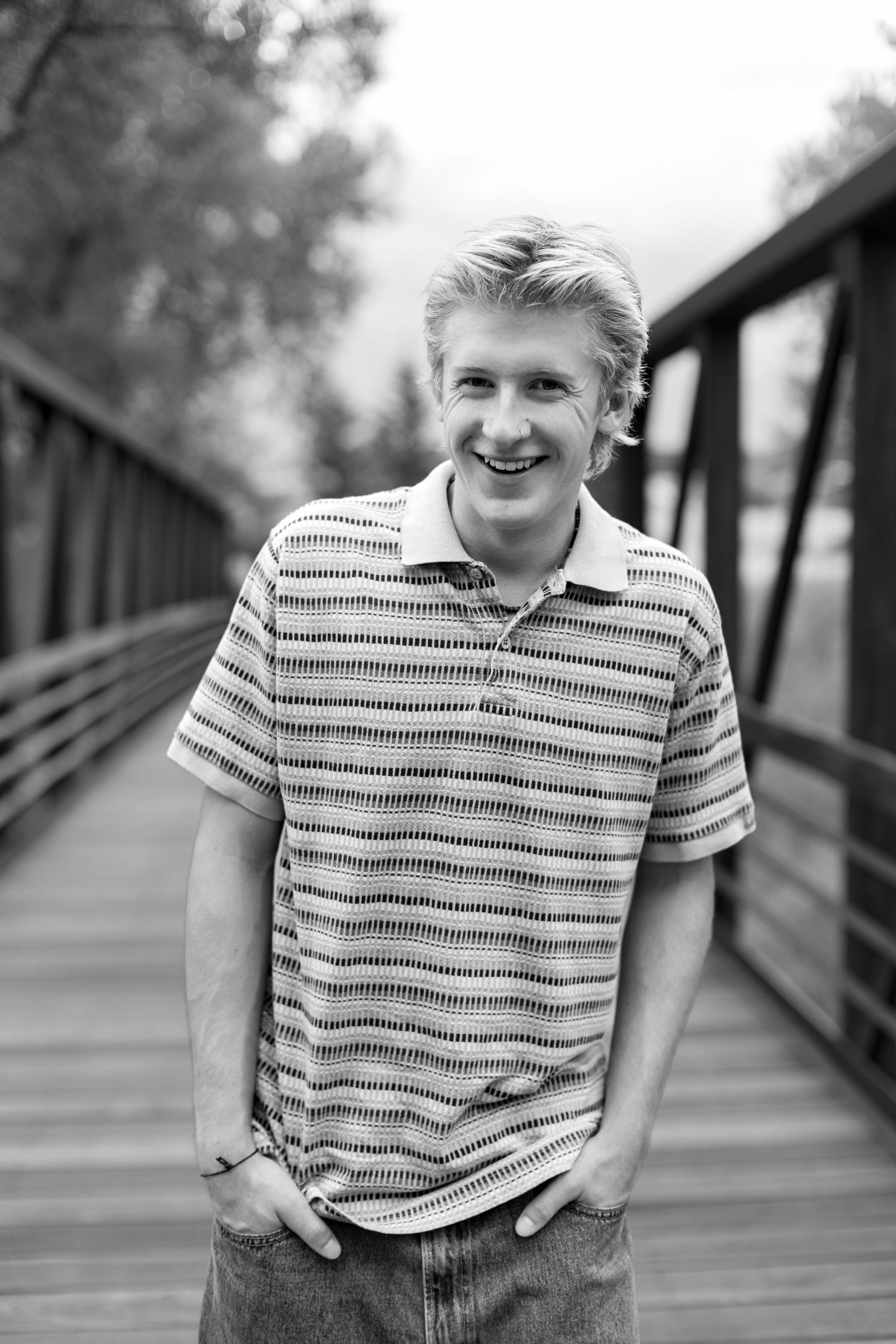Smiling teenage boy with blonde hair wearing a striped polo shirt, standing on a wooden bridge in a black and white photo.