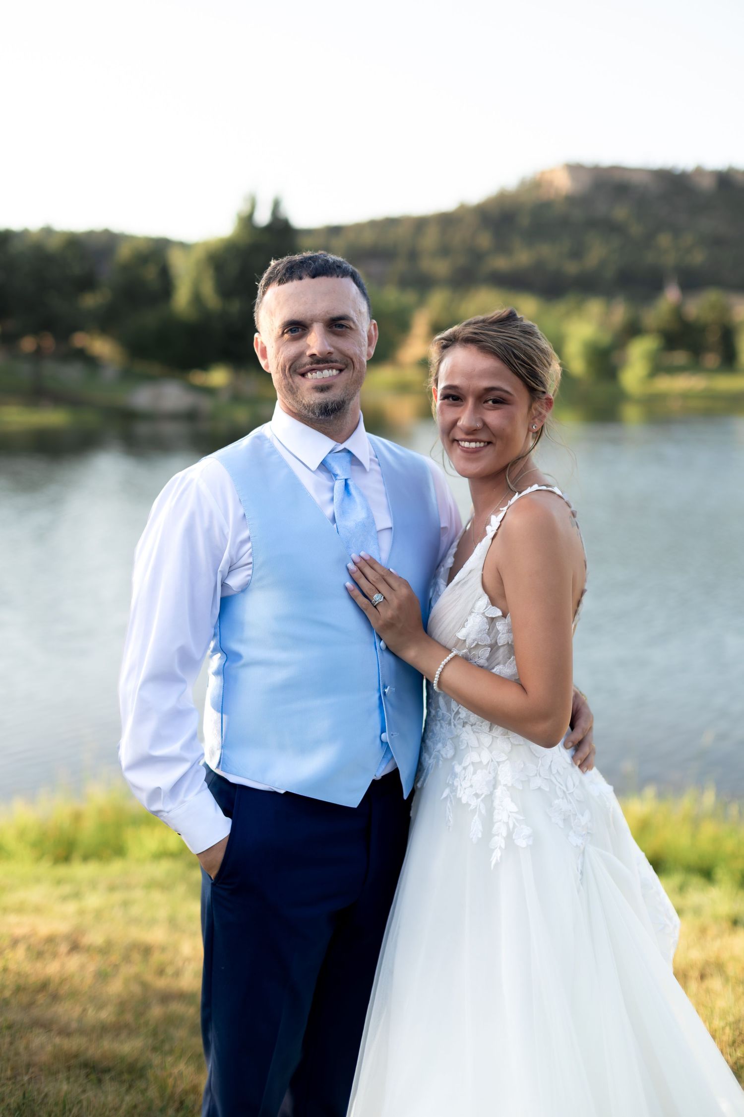 Smiling bride and groom pose together near a scenic lake with mountains in the background on their wedding day.