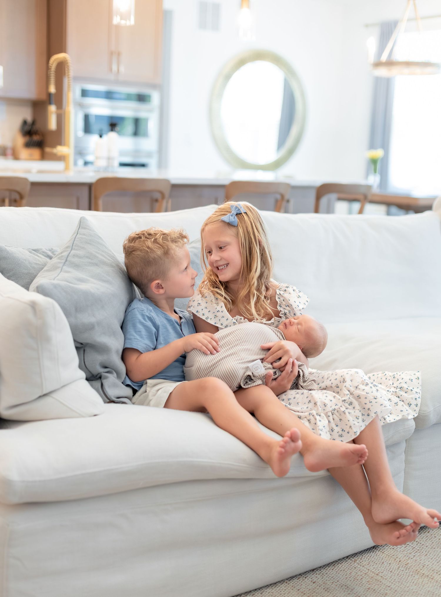 Two young siblings sit on a white couch holding a newborn baby in a bright modern home.