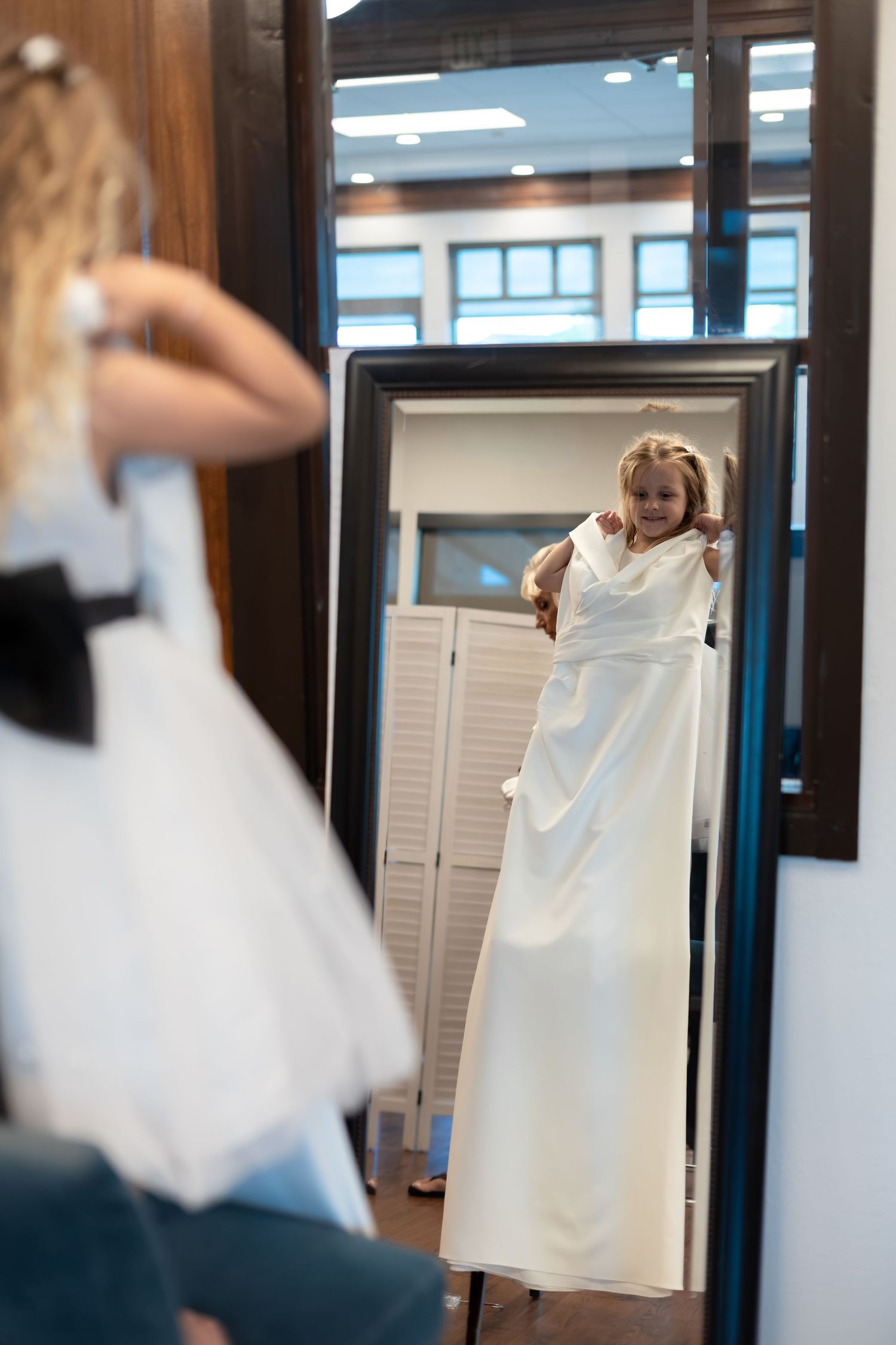 Young girl in a bridal boutique holding up a white wedding dress while looking at herself in a large mirror.