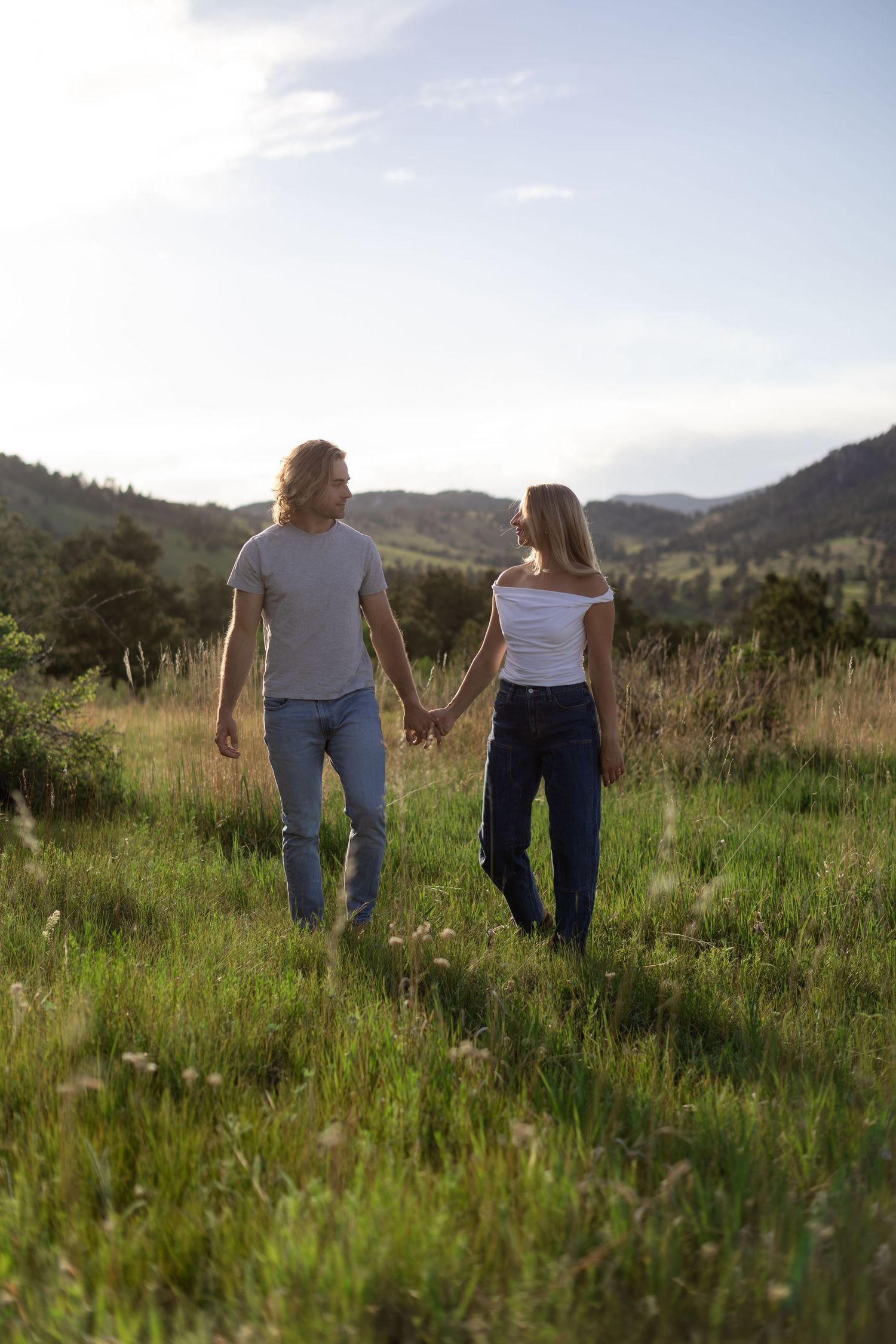 Couple holding hands walking through a lush green meadow with rolling hills and a blue sky backdrop.