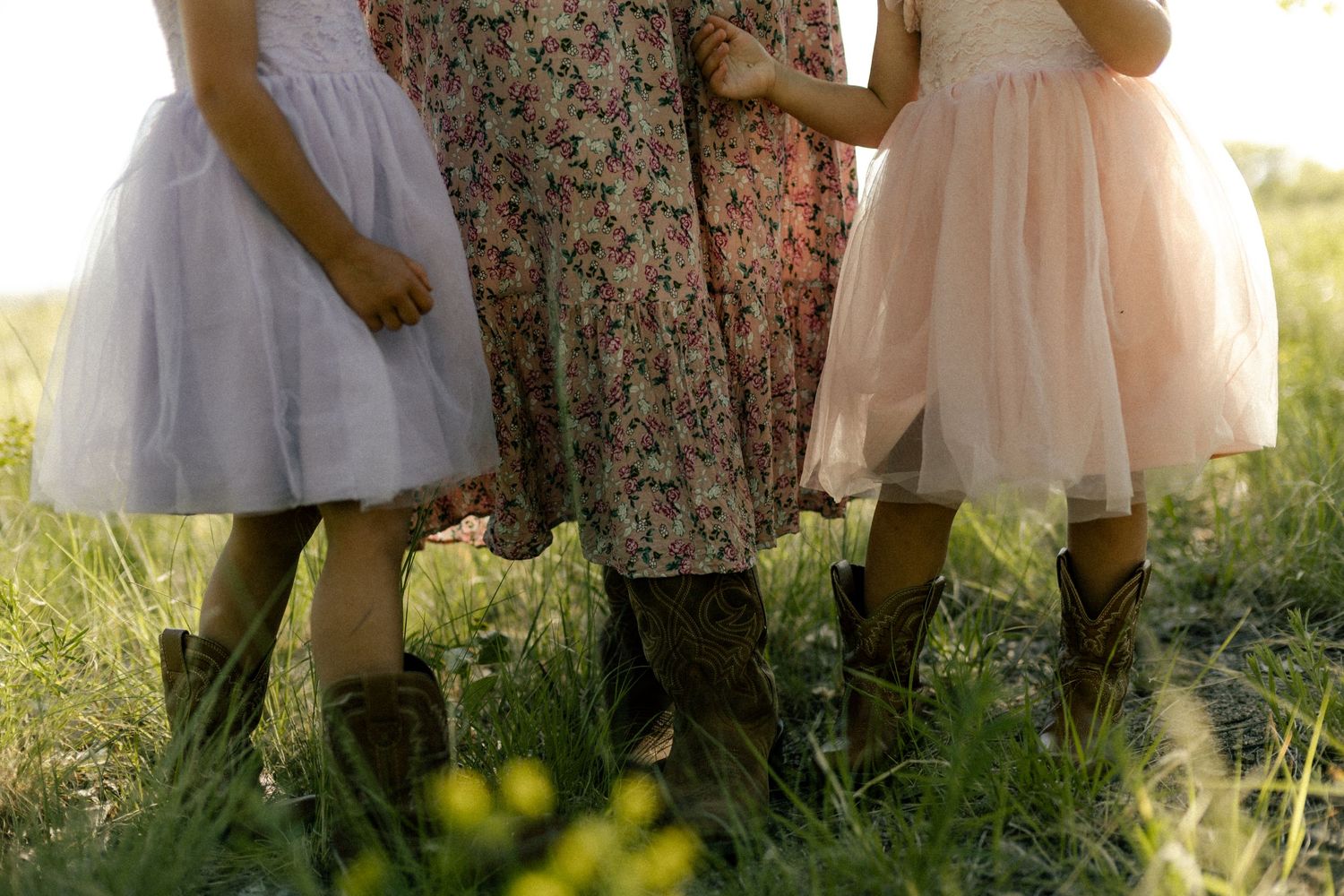 Three girls in floral and tulle dresses with cowboy boots standing in a sunny green field.
