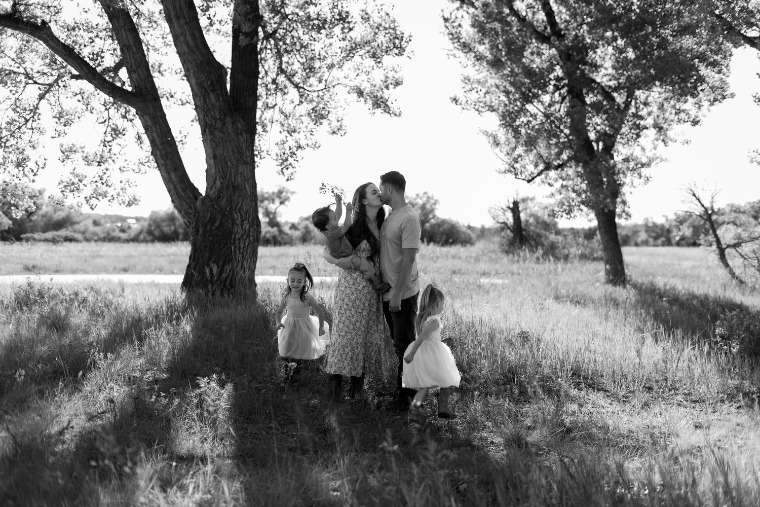Black and white family photo under trees in a field, parents kissing while children gather around them.