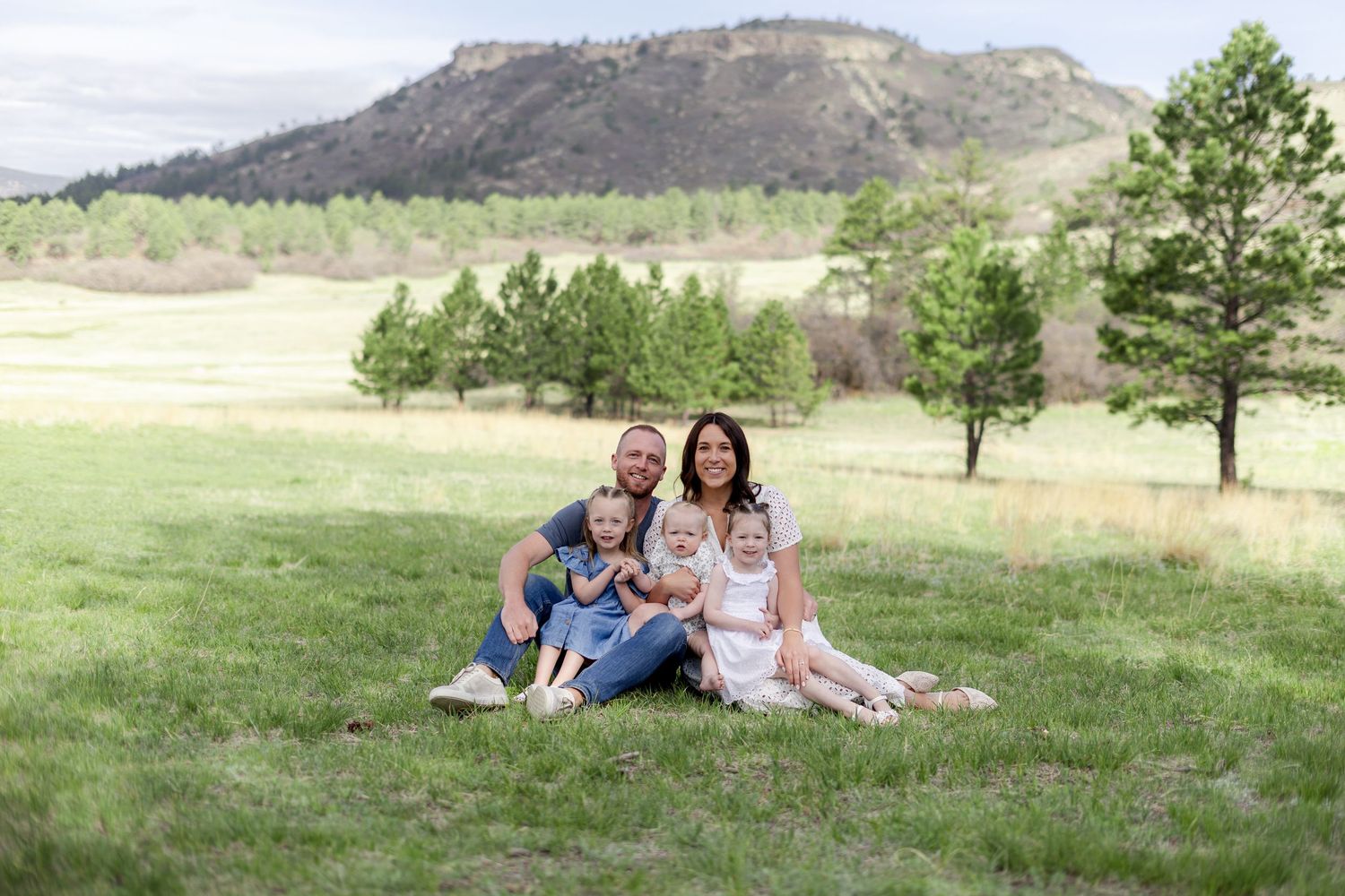 Family of four smiling together on green grass with scenic mountain and pine trees in background.