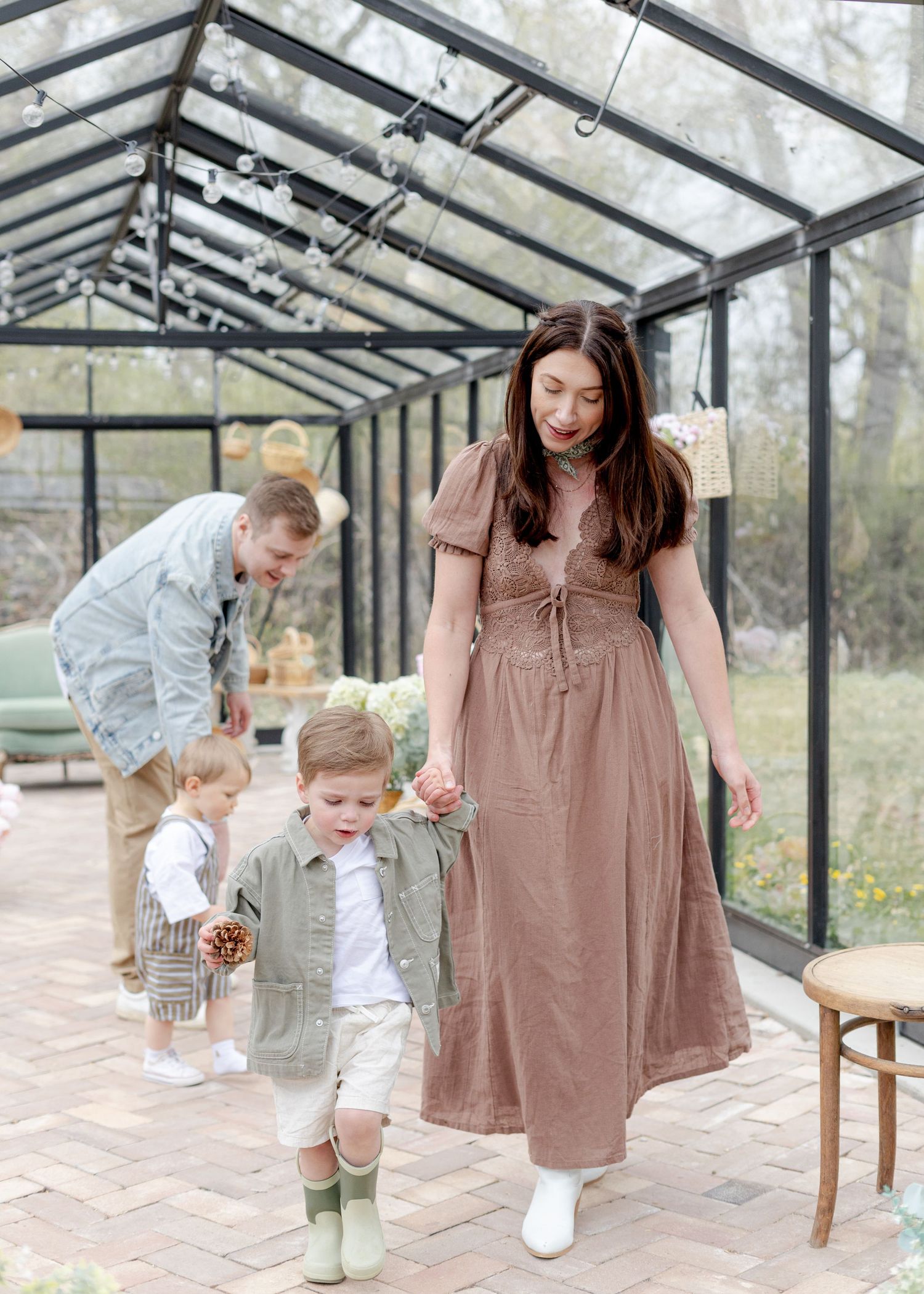 A woman in a mauve midi dress walks with a toddler in a greenhouse while a man tends to another child behind them.
