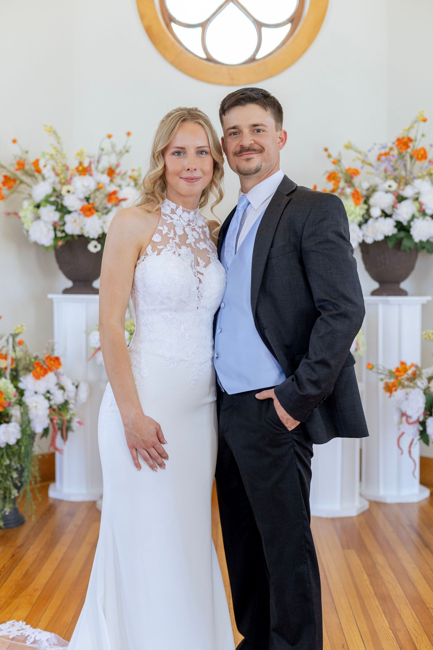Bride in white lace halter gown and groom in dark suit pose together at floral wedding altar.