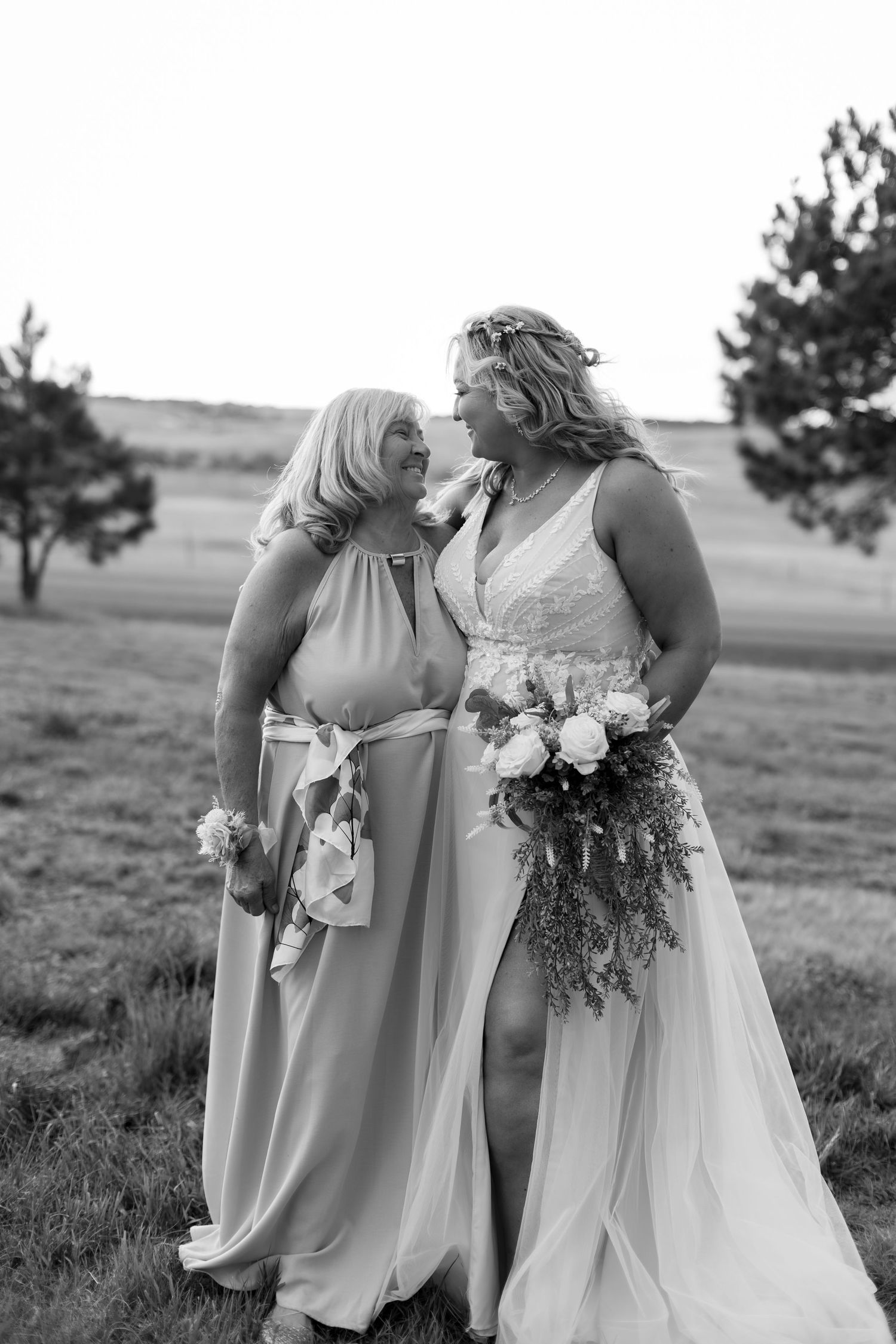 Bride in lace gown holding bouquet smiles with bridesmaid in flowing dress in outdoor field setting.