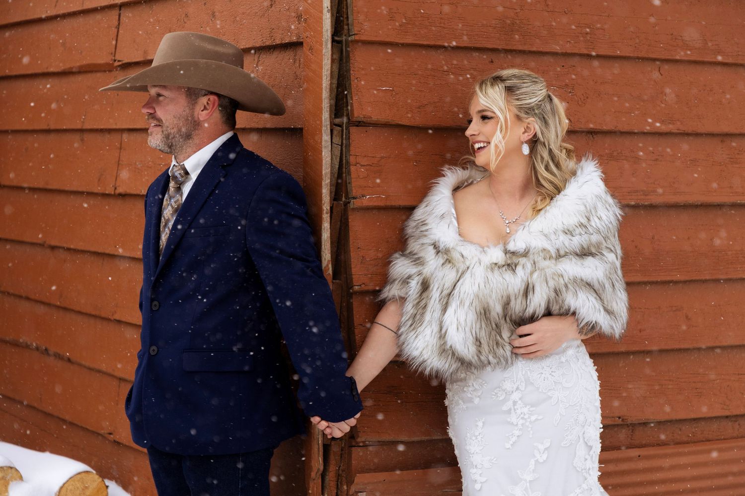 A couple in winter wedding attire shares a romantic moment against a rustic wooden wall backdrop.