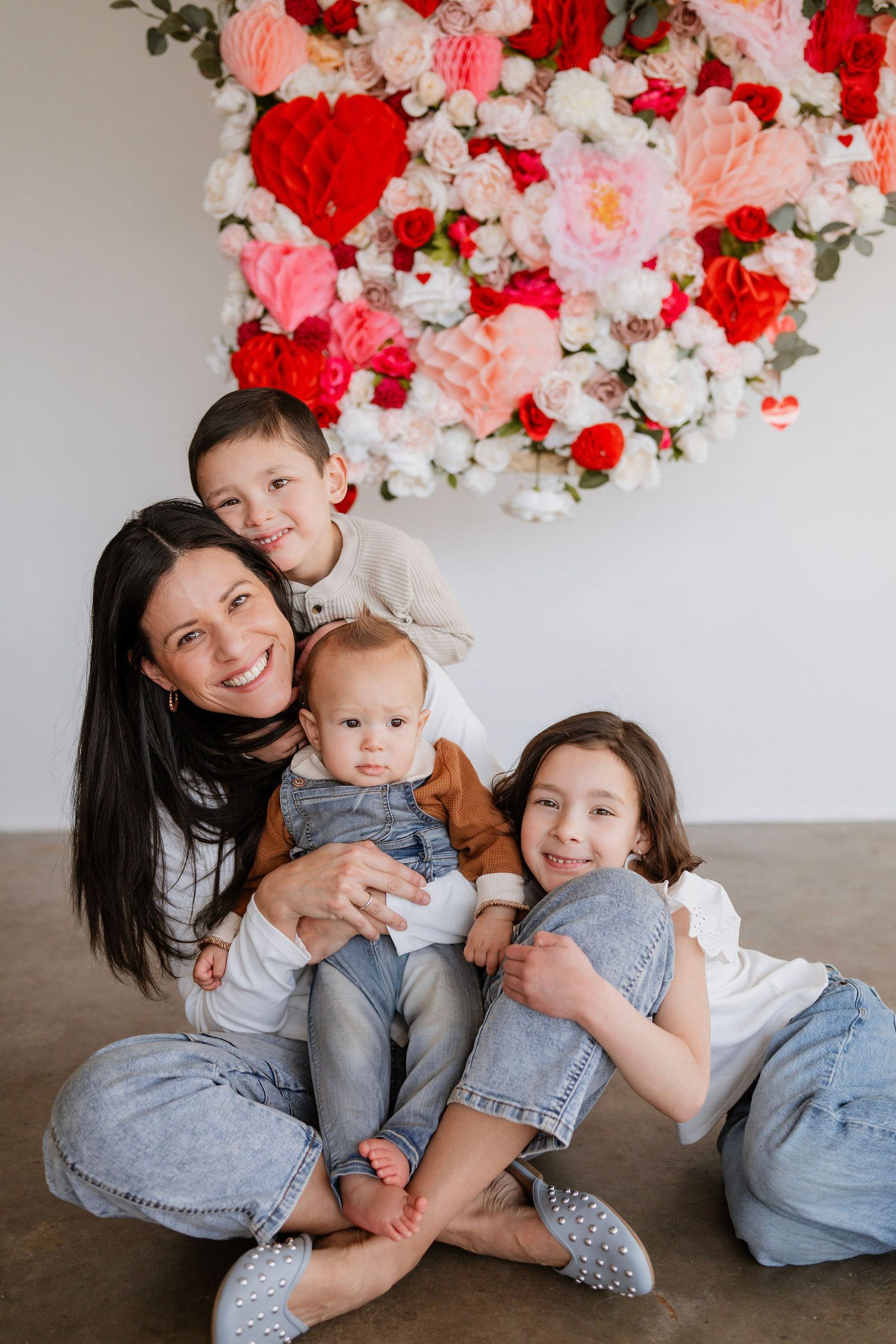 A family group sits together against a white wall decorated with red and pink floral arrangements above.