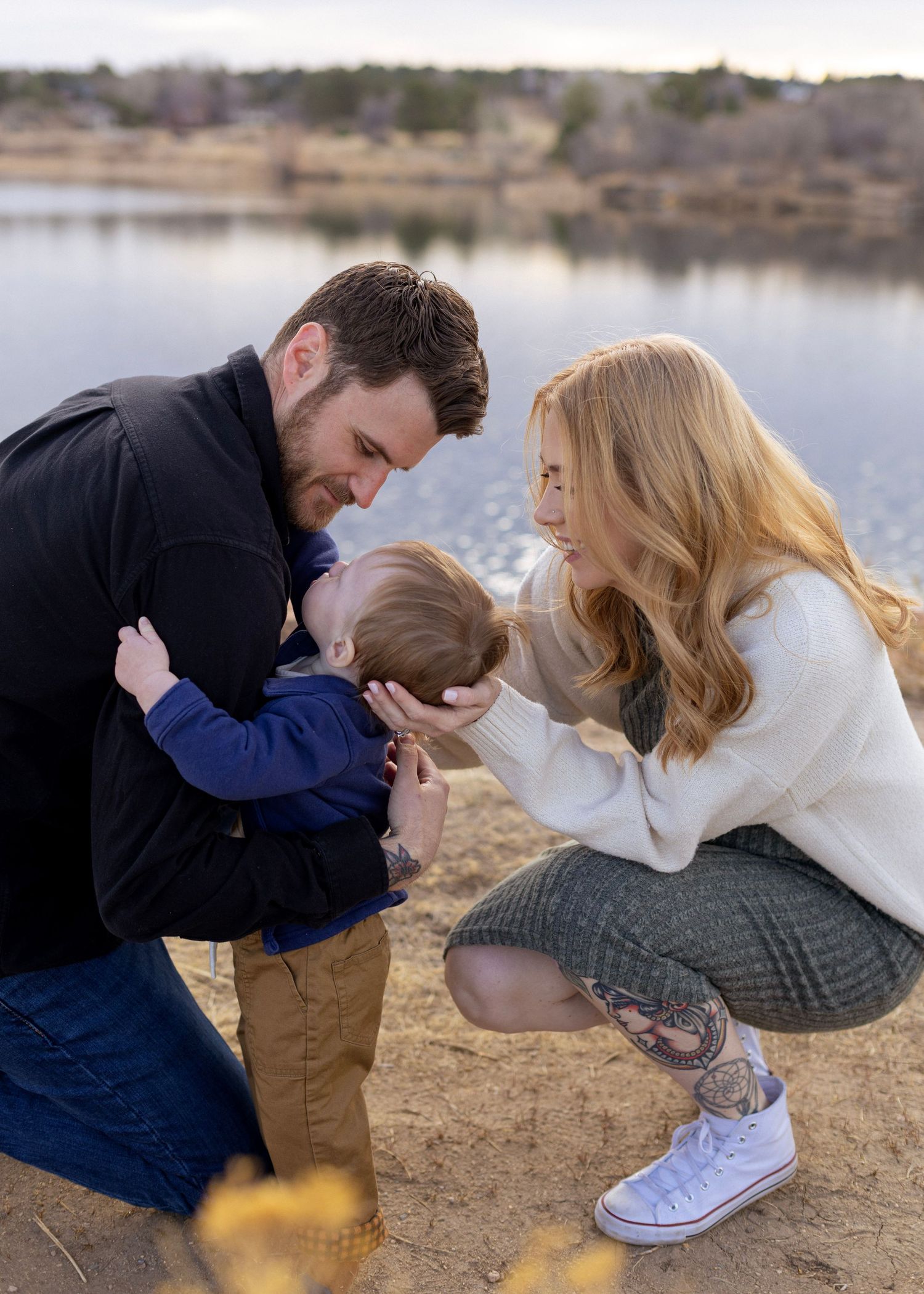 A couple shares a tender moment by a lake, sitting on sandy ground in coordinated navy and white outfits.
