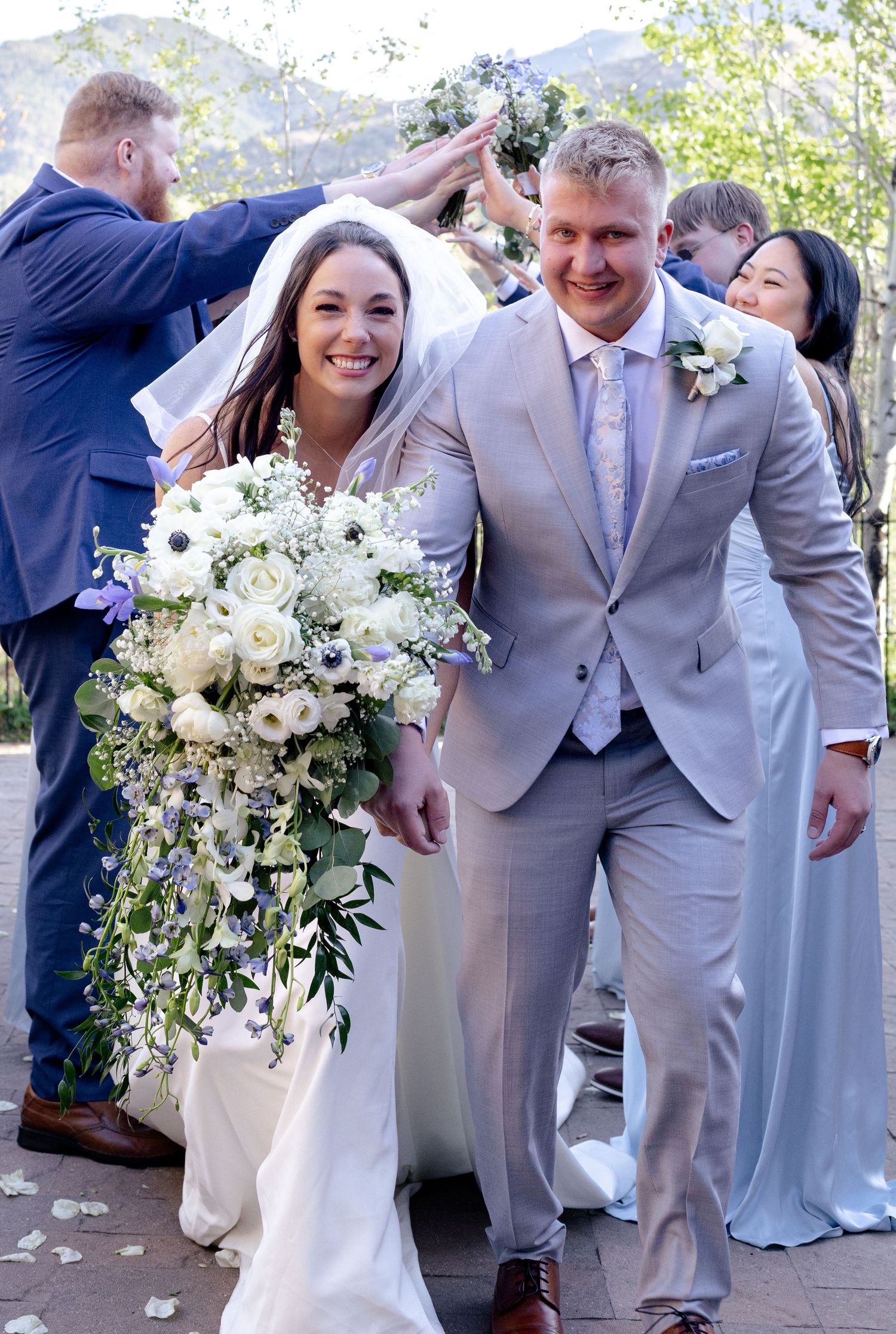 Smiling bride and groom exit wedding ceremony as guests celebrate with raised hands outdoors.