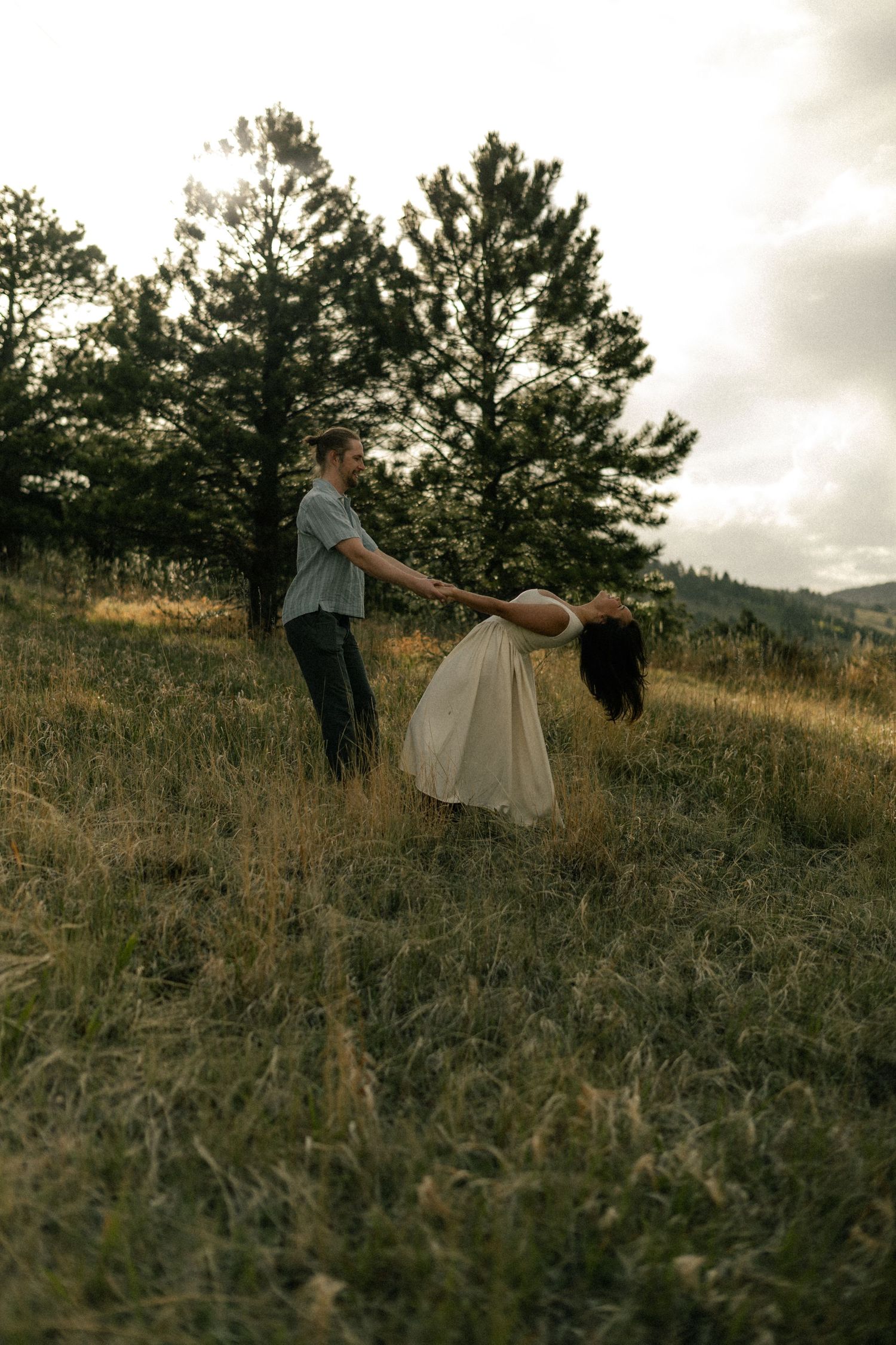 A couple dances in a grassy meadow, man spinning woman in cream dress, pine trees and cloudy sky behind them.