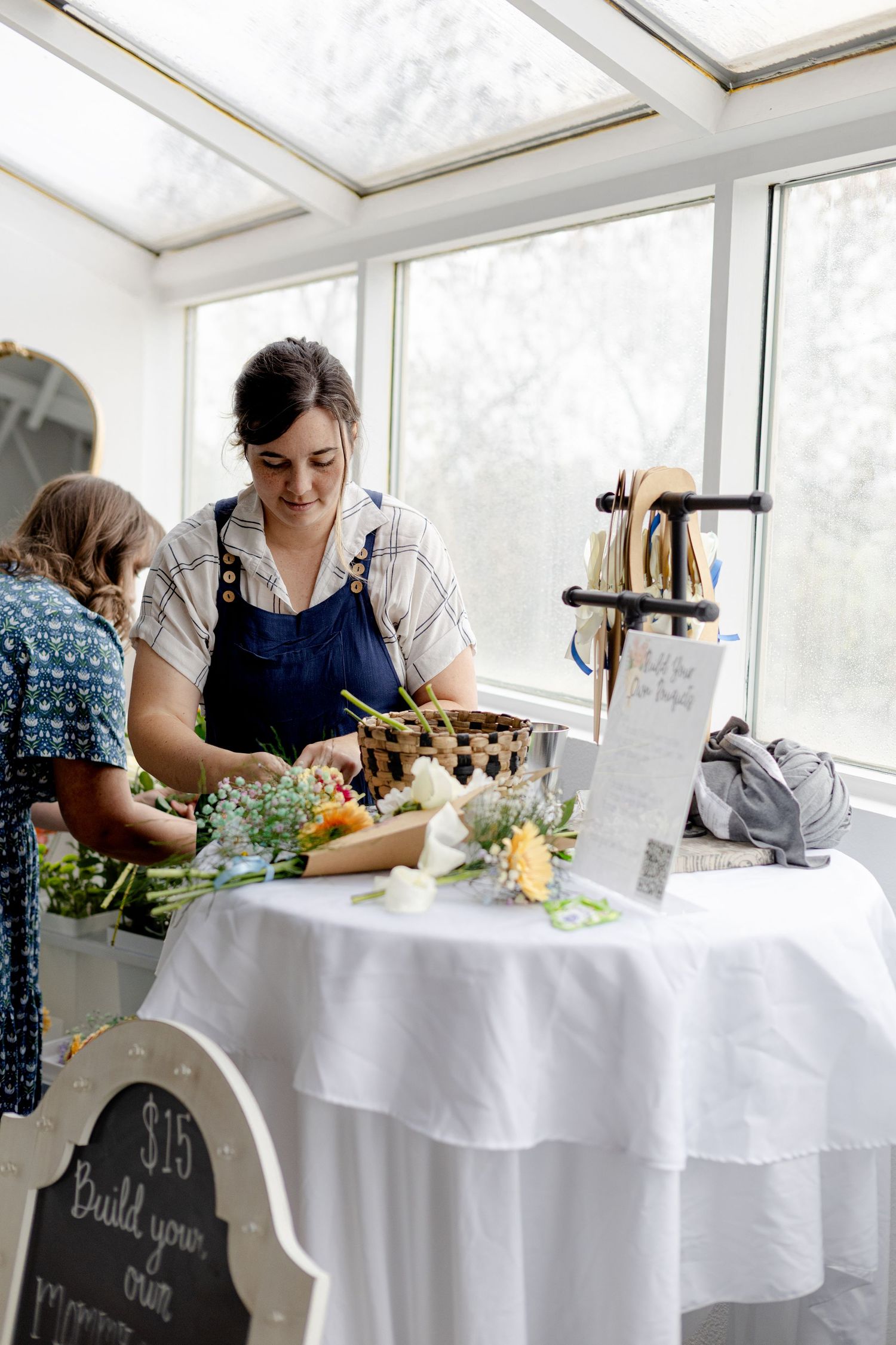 Woman in apron arranging flowers at a white tablecloth market booth with a $15 build your own bouquet sign.