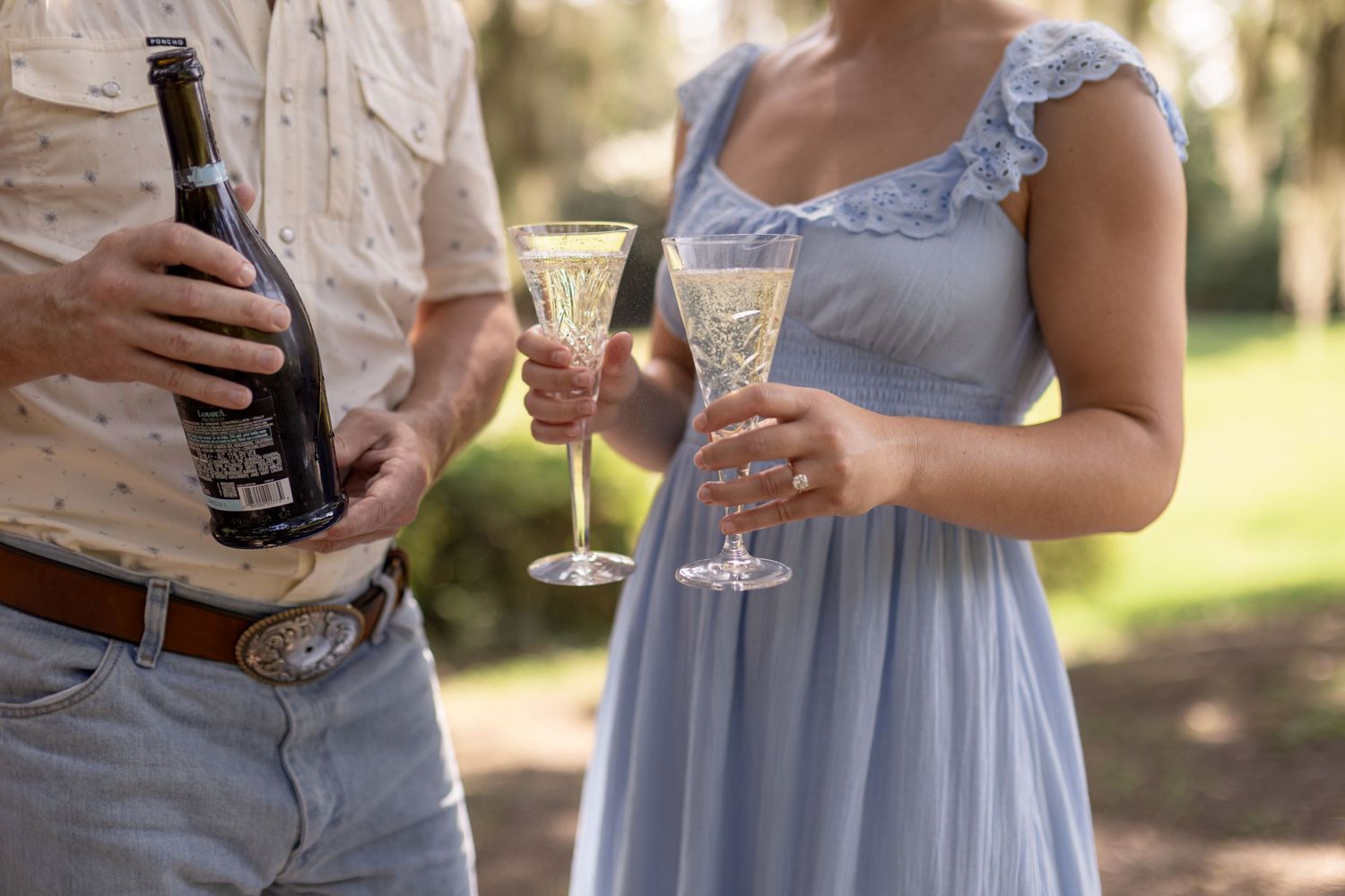 Couple celebrating outdoors, man holding wine bottle, woman in blue gown holding crystal champagne flutes at elegant event.