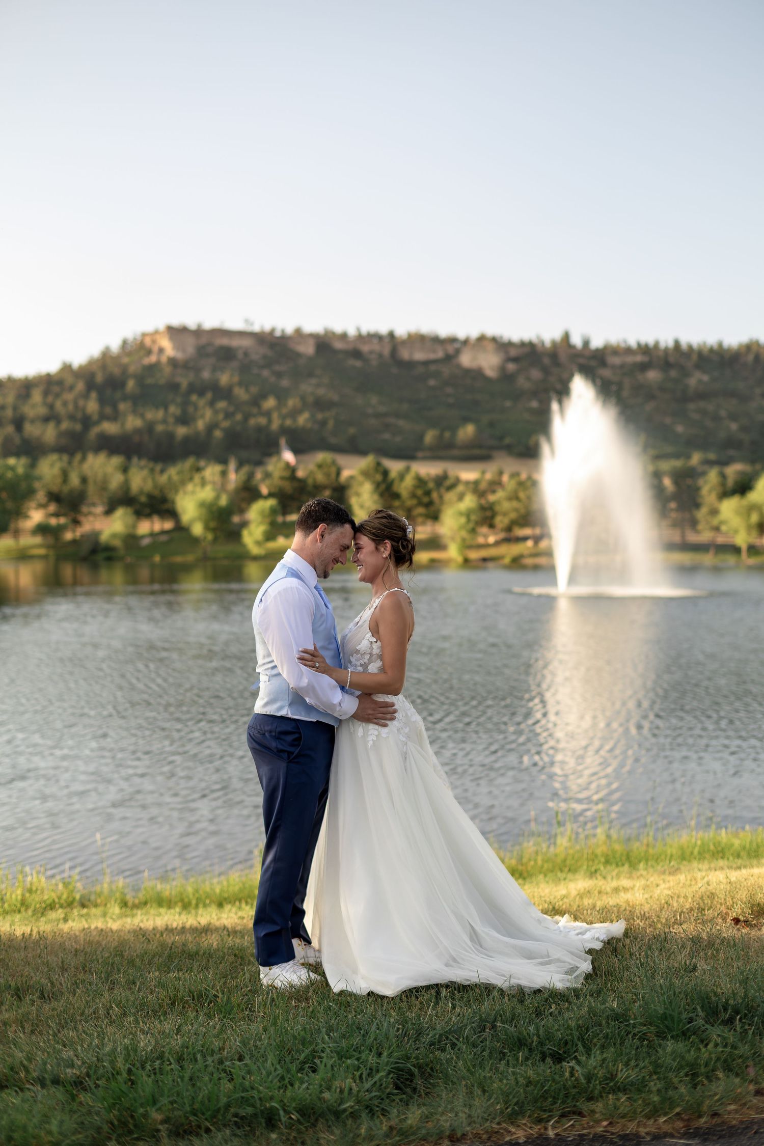 Bride and groom embrace romantically by a lake with a fountain at golden hour during an outdoor wedding.