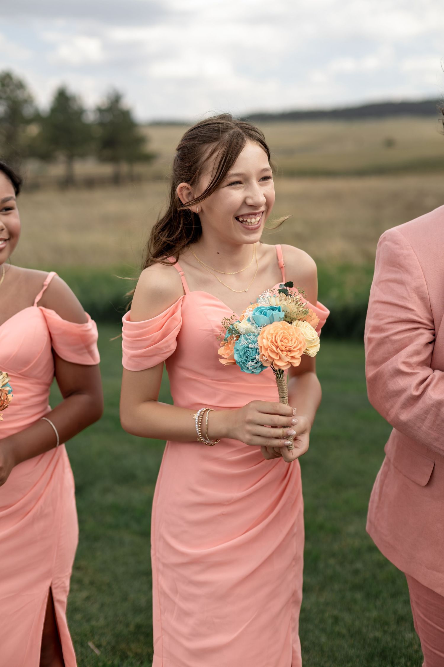 Smiling bridesmaid in coral off-shoulder dress holds colorful bouquet outdoors at wedding.